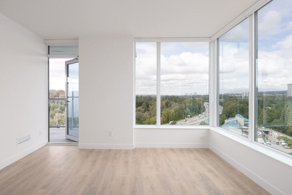 A bright, empty room with large windows and a view of a busy highway, trees, and distant buildings. The room has light wood flooring and a glass door leading to a small balcony.