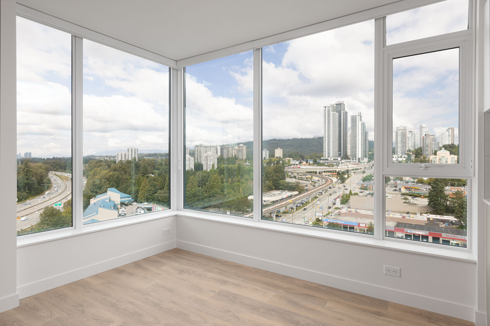 Corner room with large windows providing a panoramic view of a cityscape, including high-rise buildings, roads, and greenery under a cloudy sky. Room has light-colored wood flooring and white walls.
