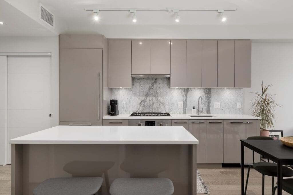 Modern kitchen with sleek gray cabinets, white countertops, marble backsplash, and stainless steel appliances. An island with two stools is in the foreground. Track lighting illuminates the space.