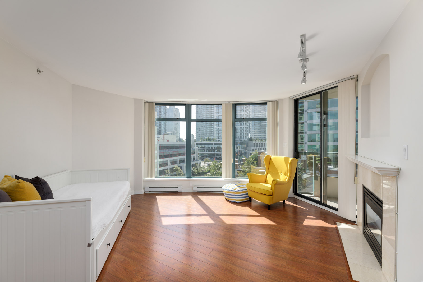 A bright living room with large windows, a yellow chair, a white daybed with cushions, wooden flooring, and a corner fireplace. Cityscape visible through the windows.