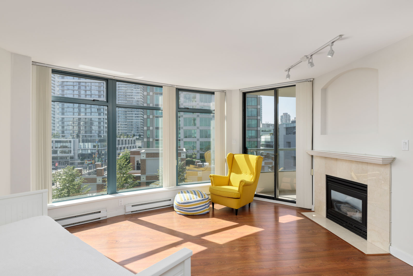 A brightly lit modern living room features a yellow armchair, a striped pouf, large windows with a city view, and a fireplace. The room has hardwood floors and track lighting.