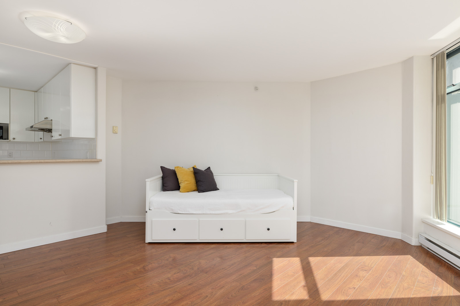 A minimalist living room with a white daybed featuring storage drawers, a couple of accent pillows, wooden flooring, and ample natural light from a large window. The adjacent kitchen area is partially visible.