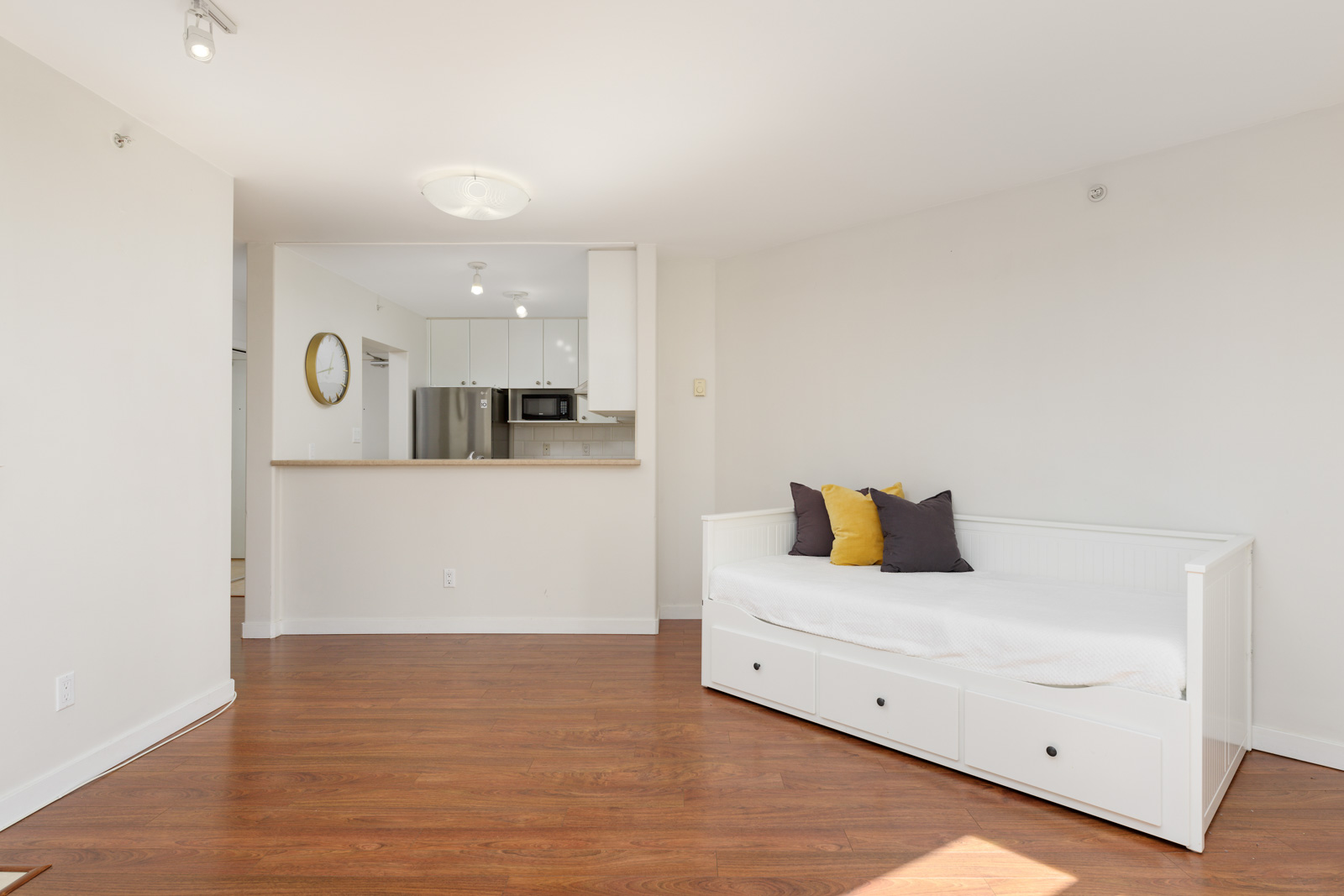 A minimalist room with white walls, wood flooring, a daybed with cushions, and a kitchen area in the background featuring white cabinetry and stainless steel appliances.