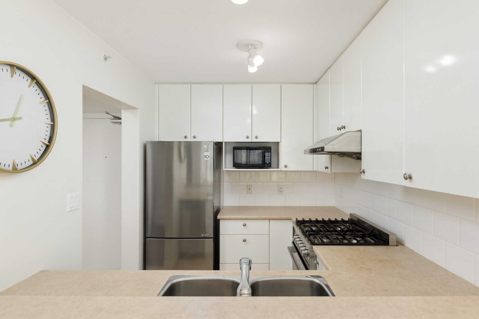 Modern kitchen with white cabinets, stainless steel refrigerator, microwave, and stove. Beige countertops, tiled backsplash, double sink, and wall clock are visible.