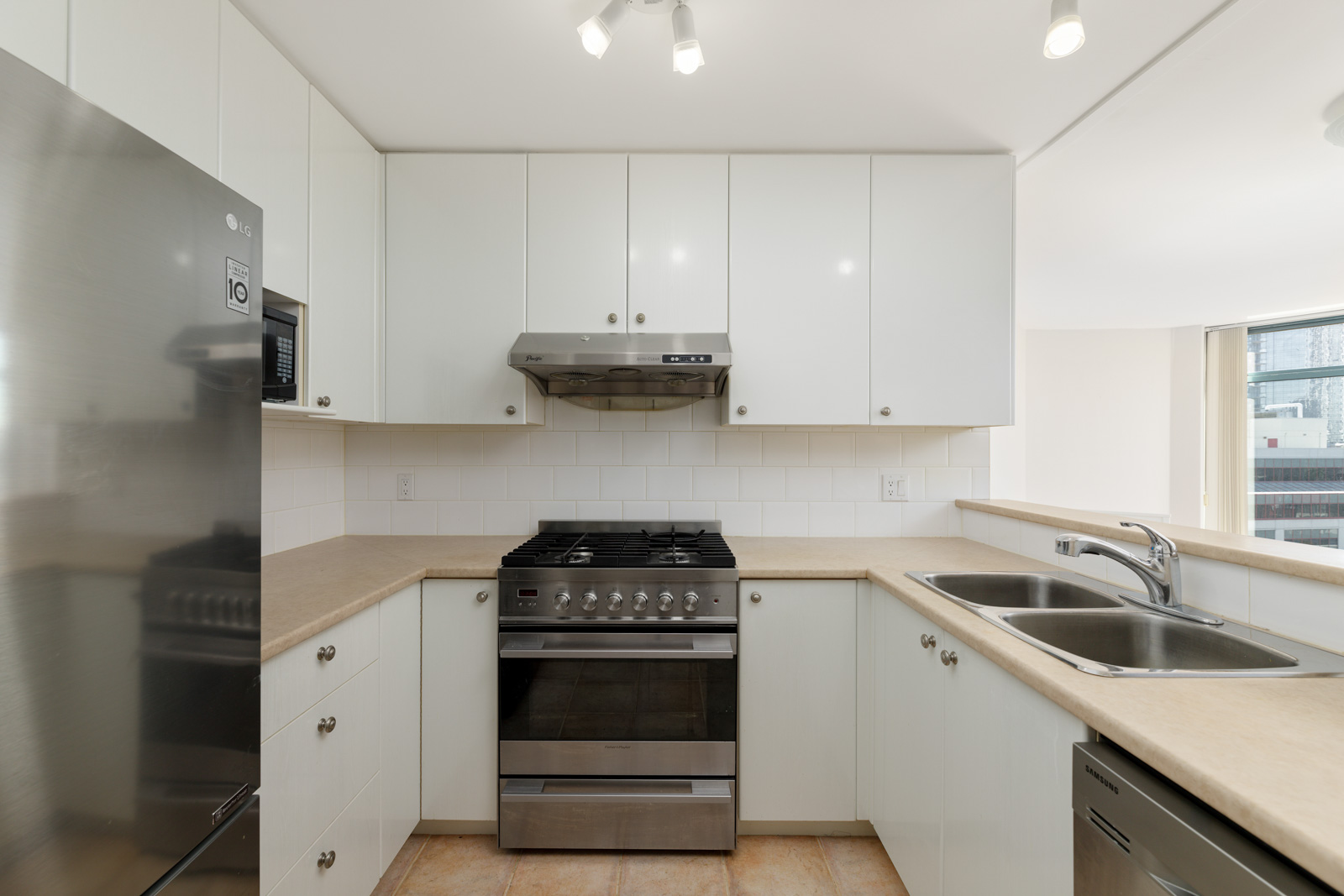 A modern kitchen featuring white cabinets, a stainless steel refrigerator, a gas stove with a range hood, tiled backsplash, dual sink, and beige countertops.
