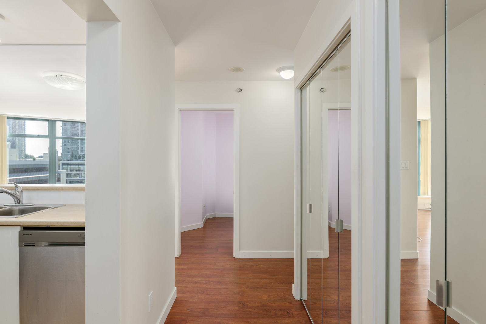A hallway with hardwood floors, mirrored closet doors, and a view into adjoining rooms and a modern kitchen area with large windows.
