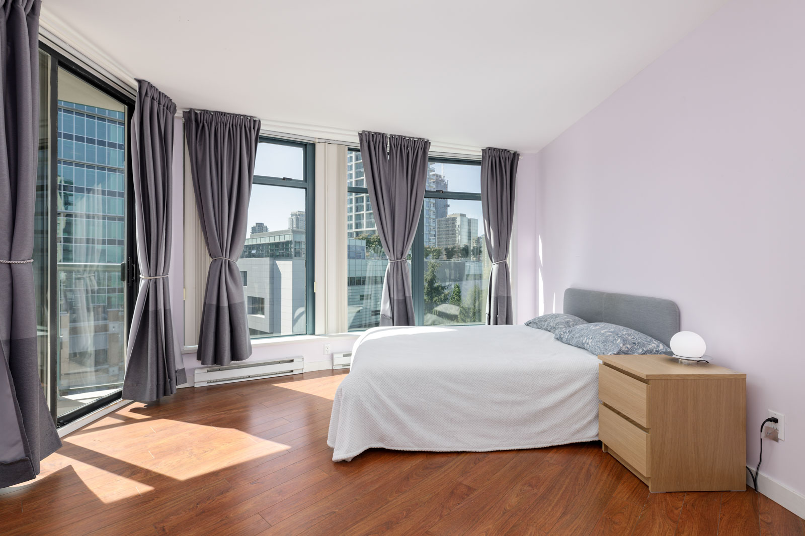 A modern bedroom with large windows covered by gray curtains, a double bed with white bedding, and a wooden nightstand with a small lamp, all set on a hardwood floor.