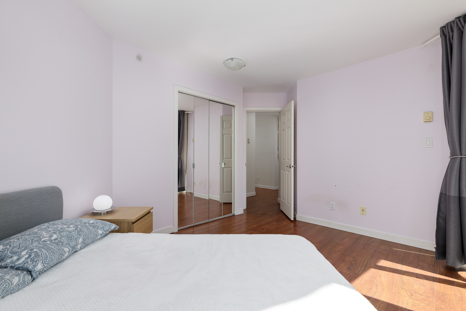 A bedroom with light purple walls, a bed, a nightstand with a lamp, mirrored closet doors, hardwood floors, and a window with gray curtains. The room is empty and well-lit.