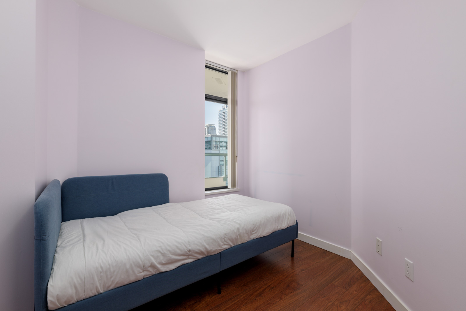 A small bedroom with light purple walls featuring a single blue bed with white bedding. There's one window showing an urban skyline and dark wooden floors.