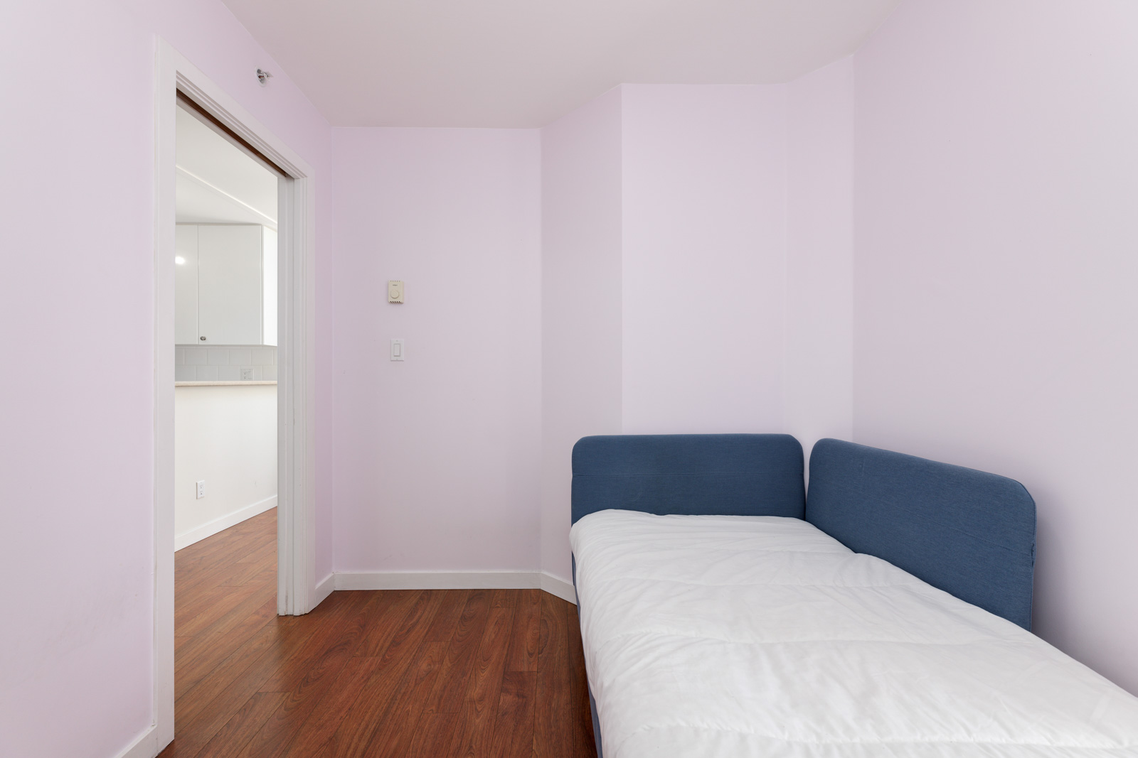 A small bedroom with light pink walls features a single blue upholstered bed and has a doorway leading to a room with wooden flooring.