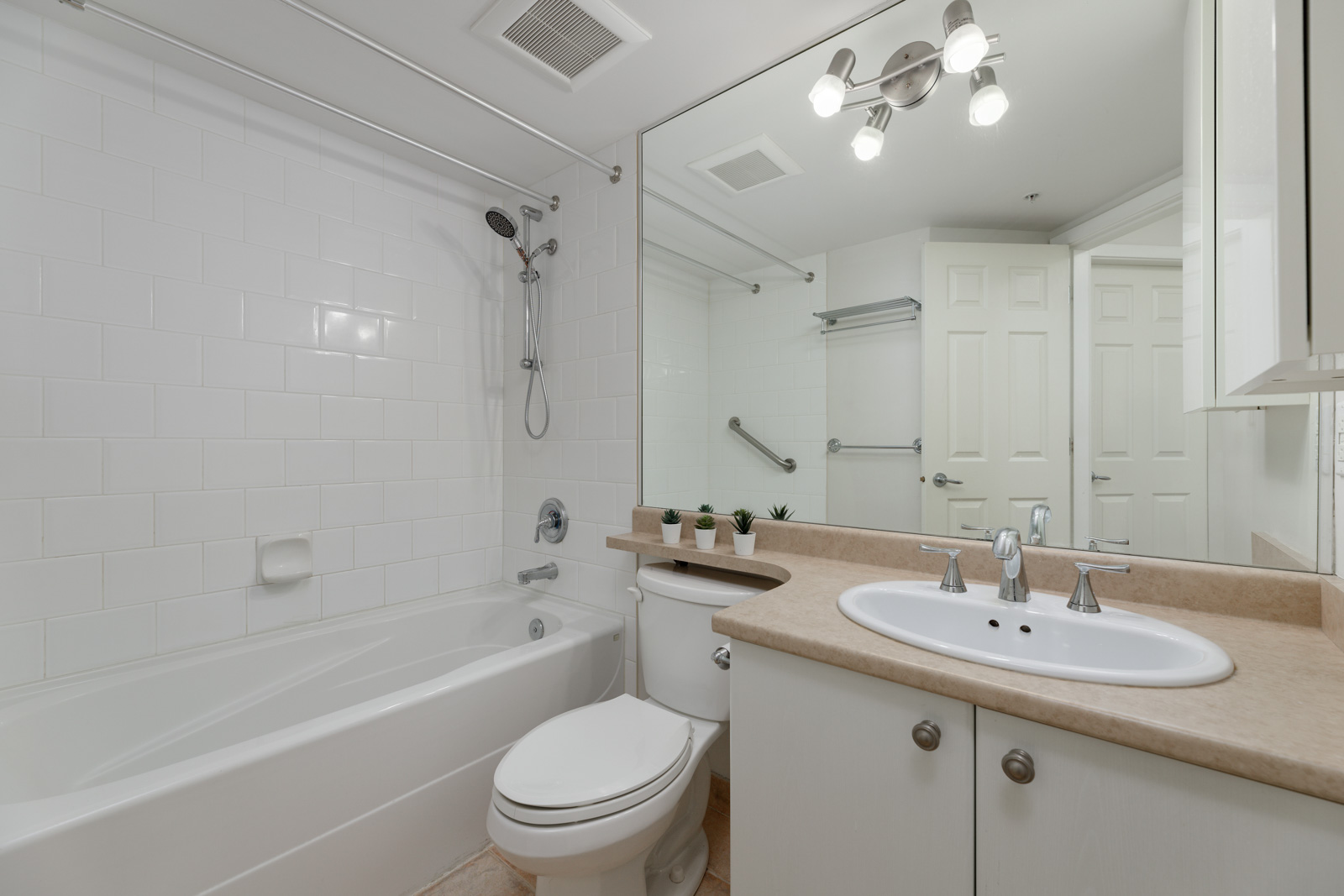 A clean, modern bathroom with a white bathtub, toilet, single sink with countertop, large mirror, and three-light fixture above. The walls are tiled, and there is a towel rack beside the door.