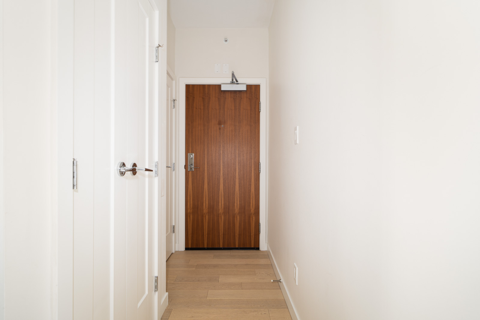 A narrow, well-lit hallway with light wooden flooring, white walls, and white doors on the left side. At the end of the hallway is a wooden door with a metal handle and a door closer at the top.