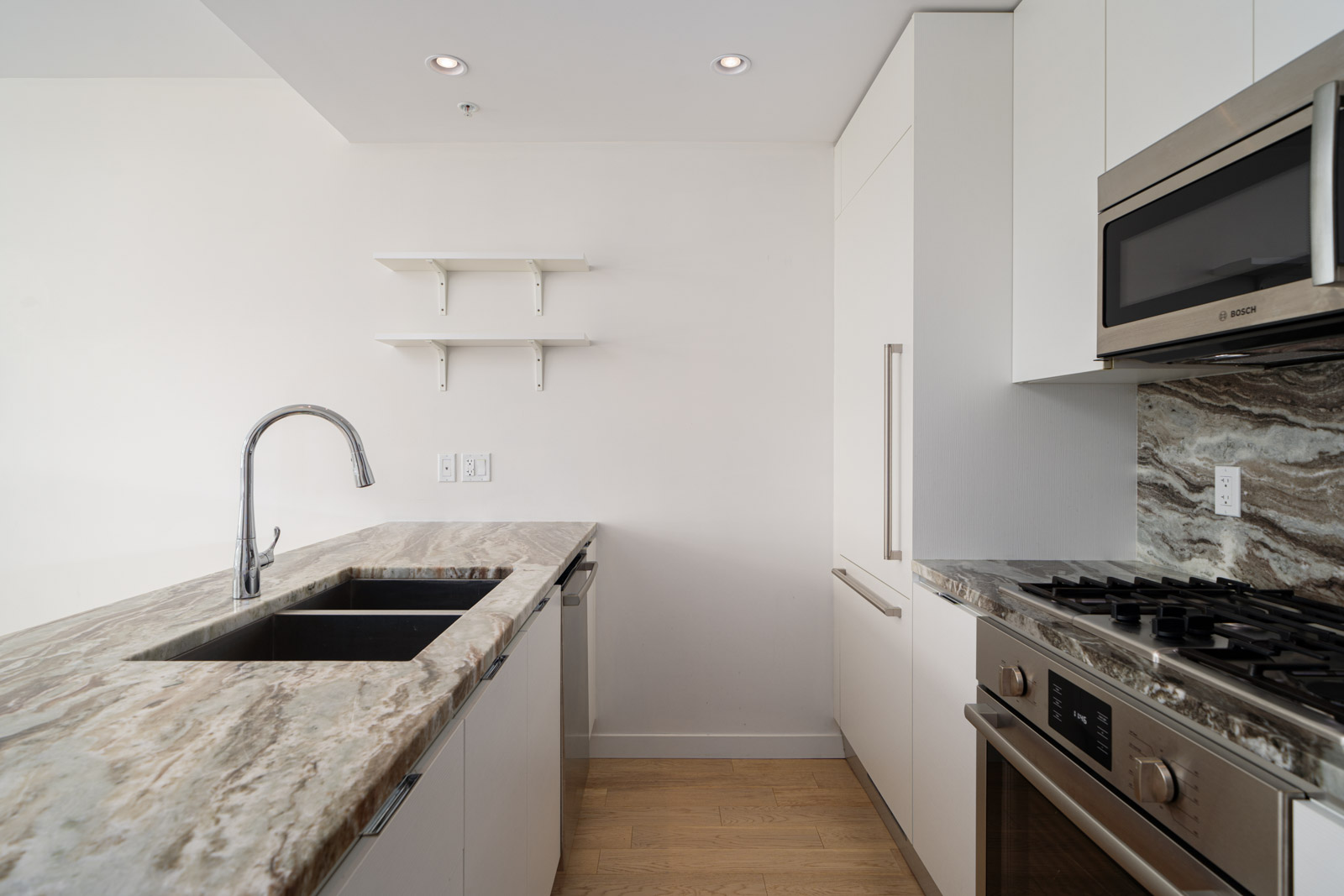 A modern kitchen features white cabinetry, stainless steel appliances, a marble countertop, a built-in microwave, a gas stove, an under-mount sink with a gooseneck faucet, and wooden flooring.