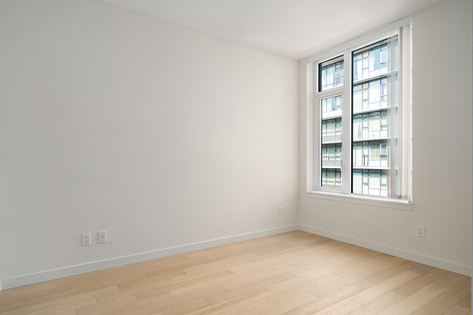 A small, empty room with light-colored wooden flooring and a large window with vertical blinds.