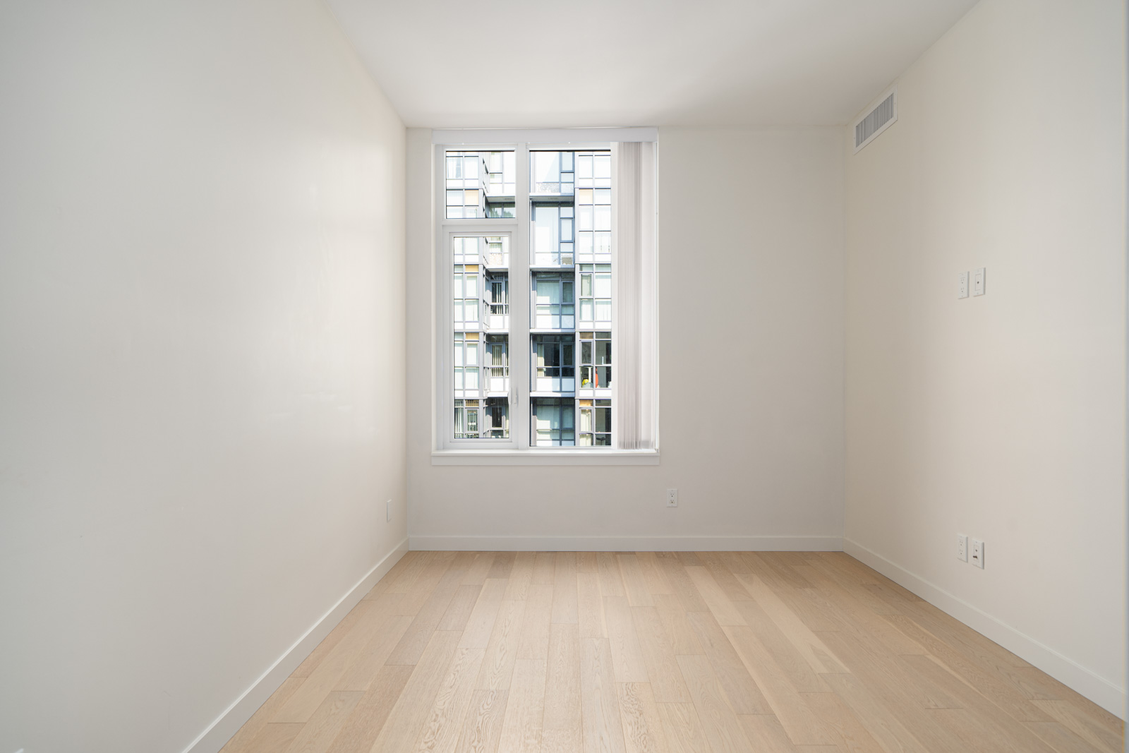 An empty room with light-colored wooden floors, white walls, and a large window showing the exterior of a modern building.