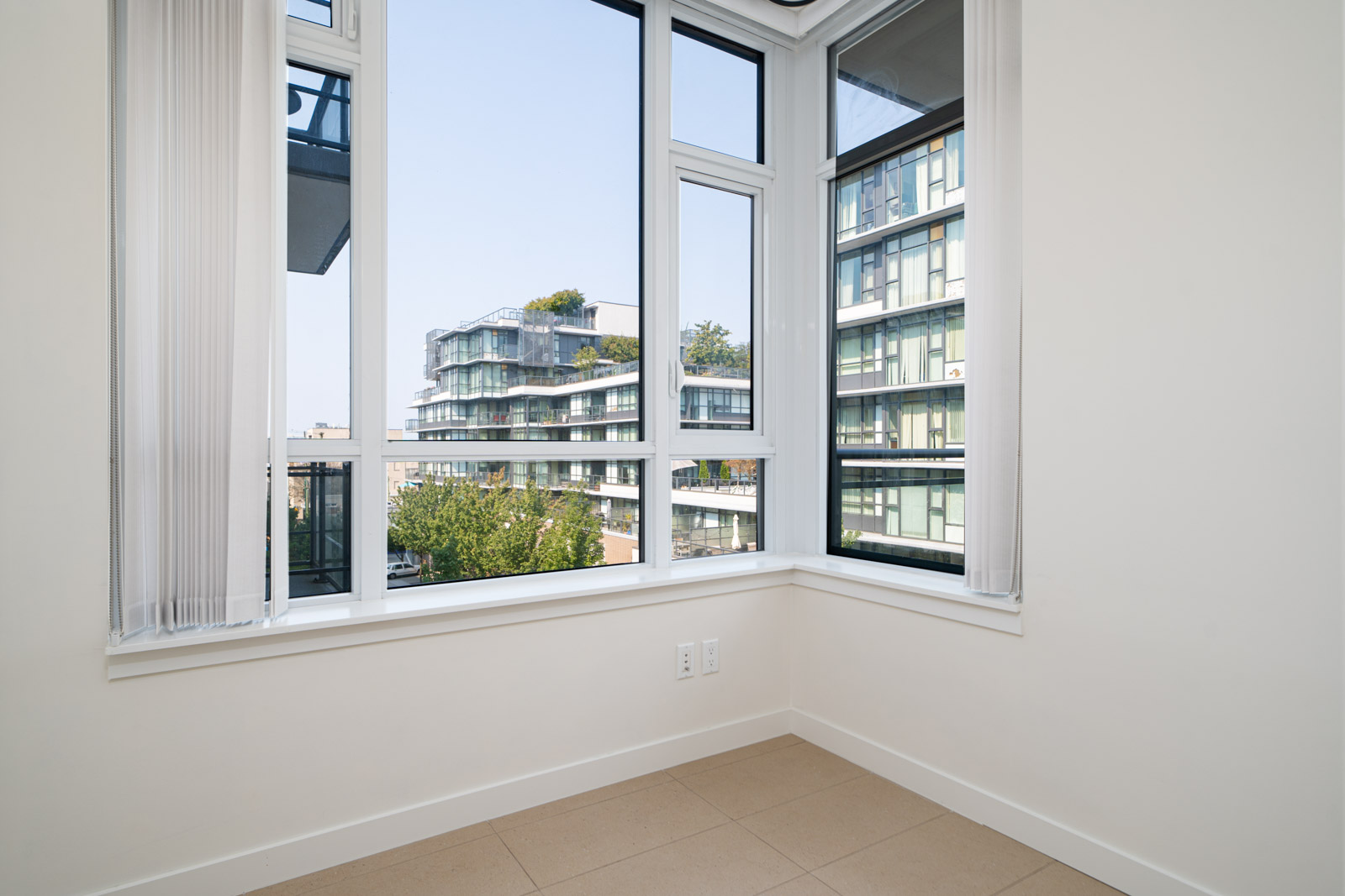 Empty room with large windows and a view of modern apartment buildings; white walls and tiled floor.