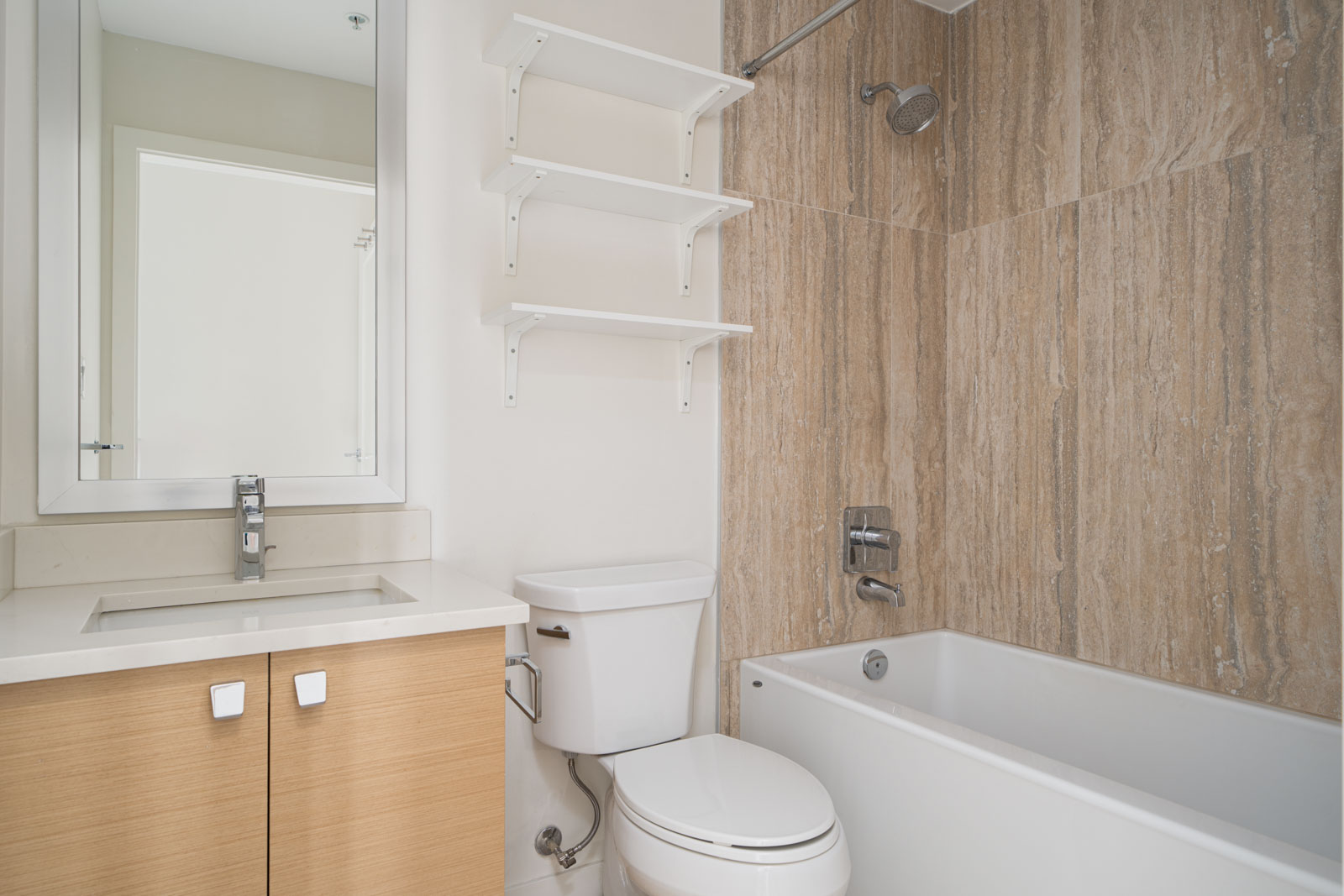 A modern bathroom with a white toilet, a sink with a wooden cabinet, a wall-mounted mirror, a bathtub with a showerhead, and beige tiled walls around the bathtub.