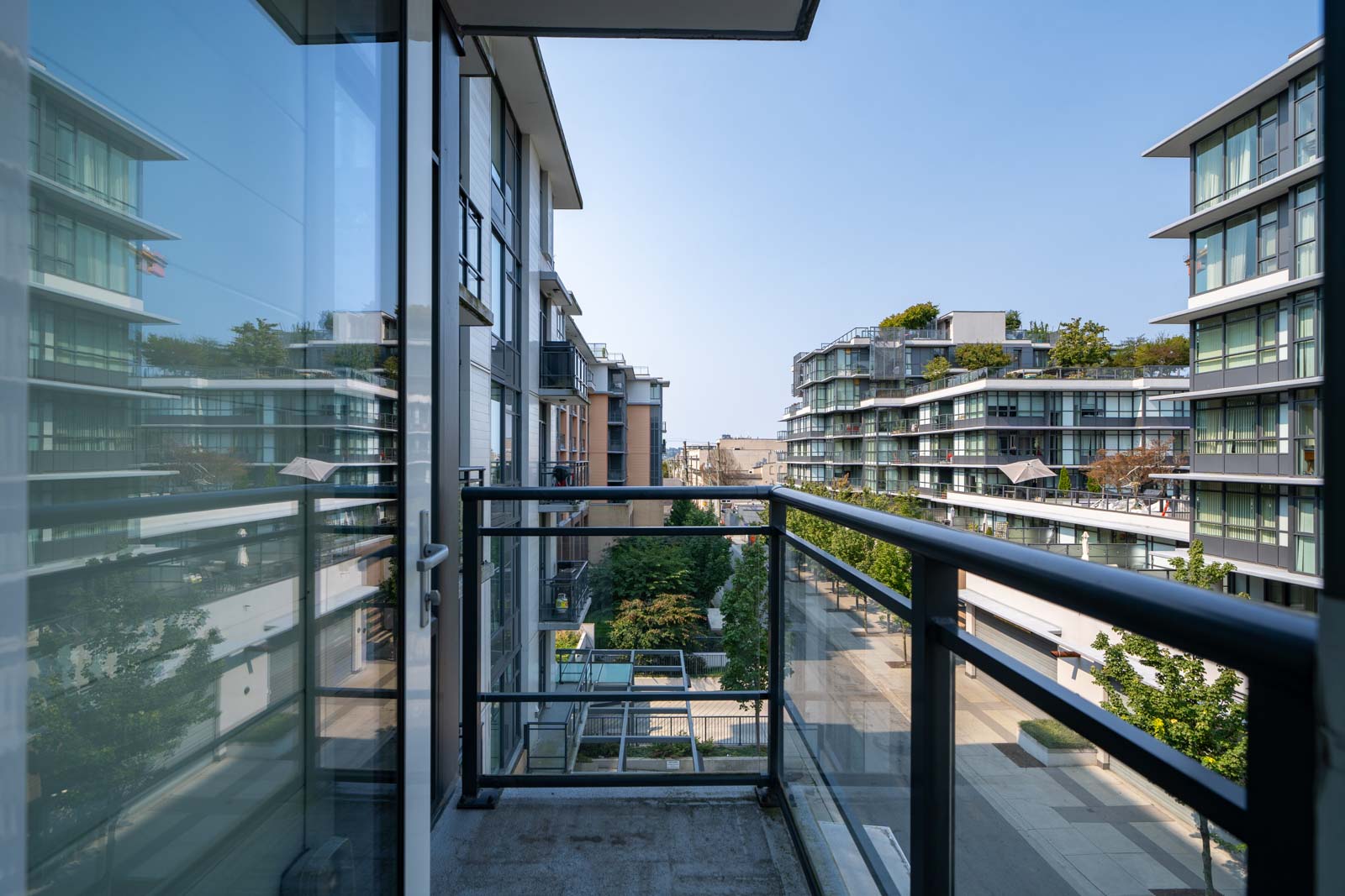 A view from a balcony showing modern, multi-story apartment buildings with glass windows and balconies, overlooking a courtyard with greenery.