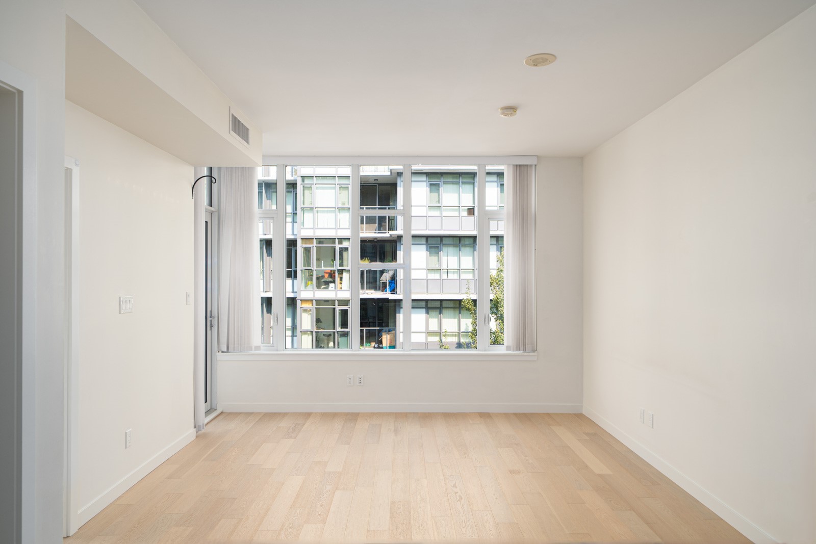 An unfurnished room with light wood flooring, white walls, and large windows offering a view of neighboring buildings.