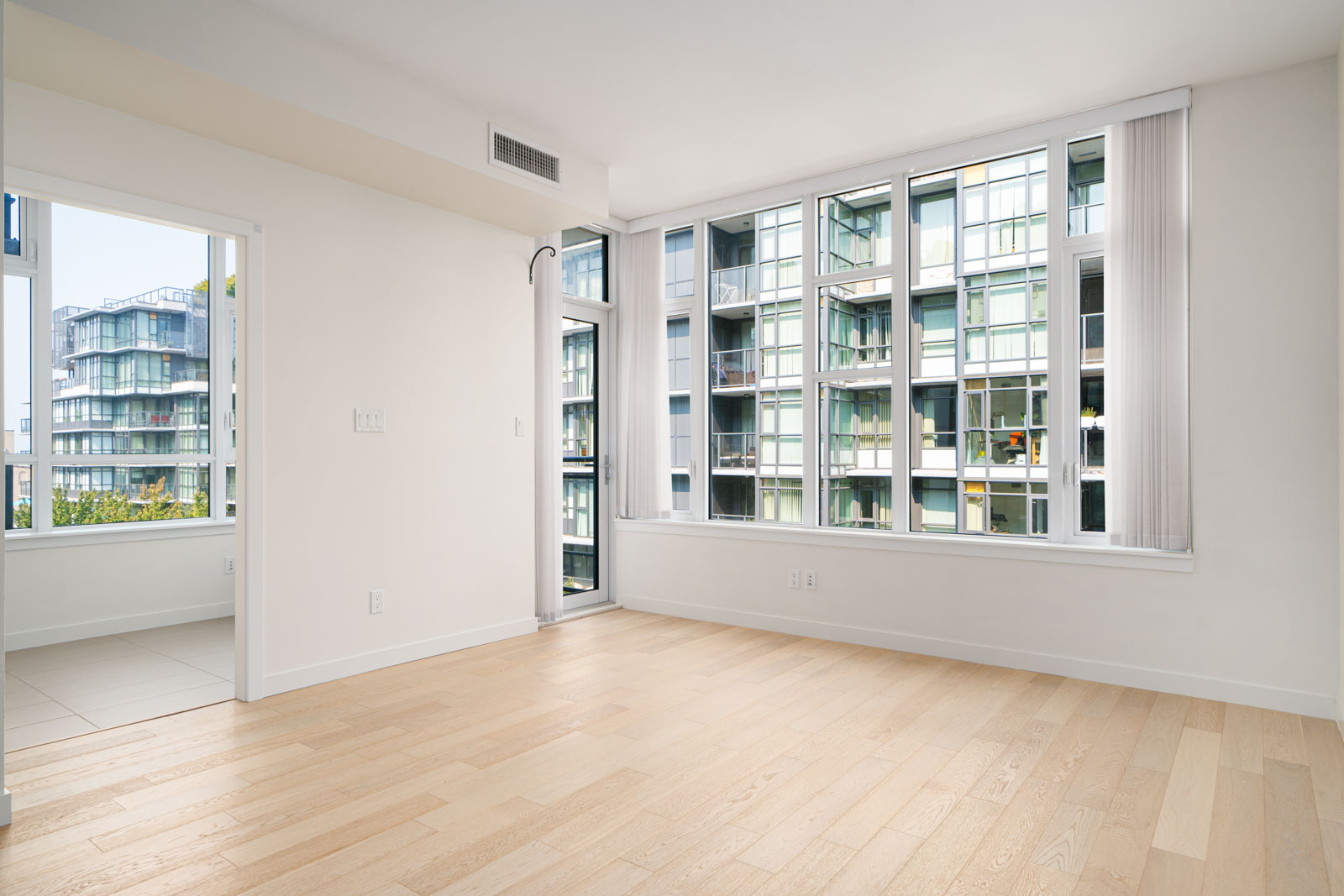 A bright, empty room with light-colored hardwood flooring, large windows, a doorway to an adjoining room, and a view of modern apartment buildings.