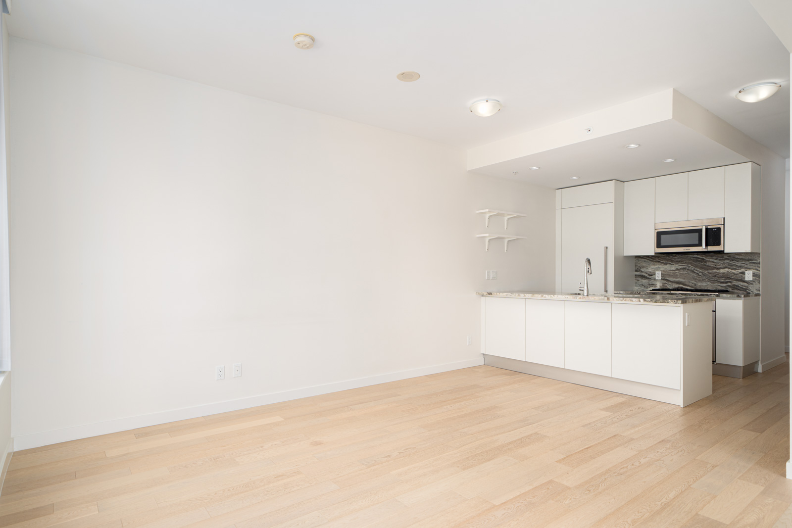 A modern apartment with light wood flooring, white walls, and a kitchen area featuring white cabinets, a marble countertop, and stainless steel appliances. The room is well-lit by ceiling lights.