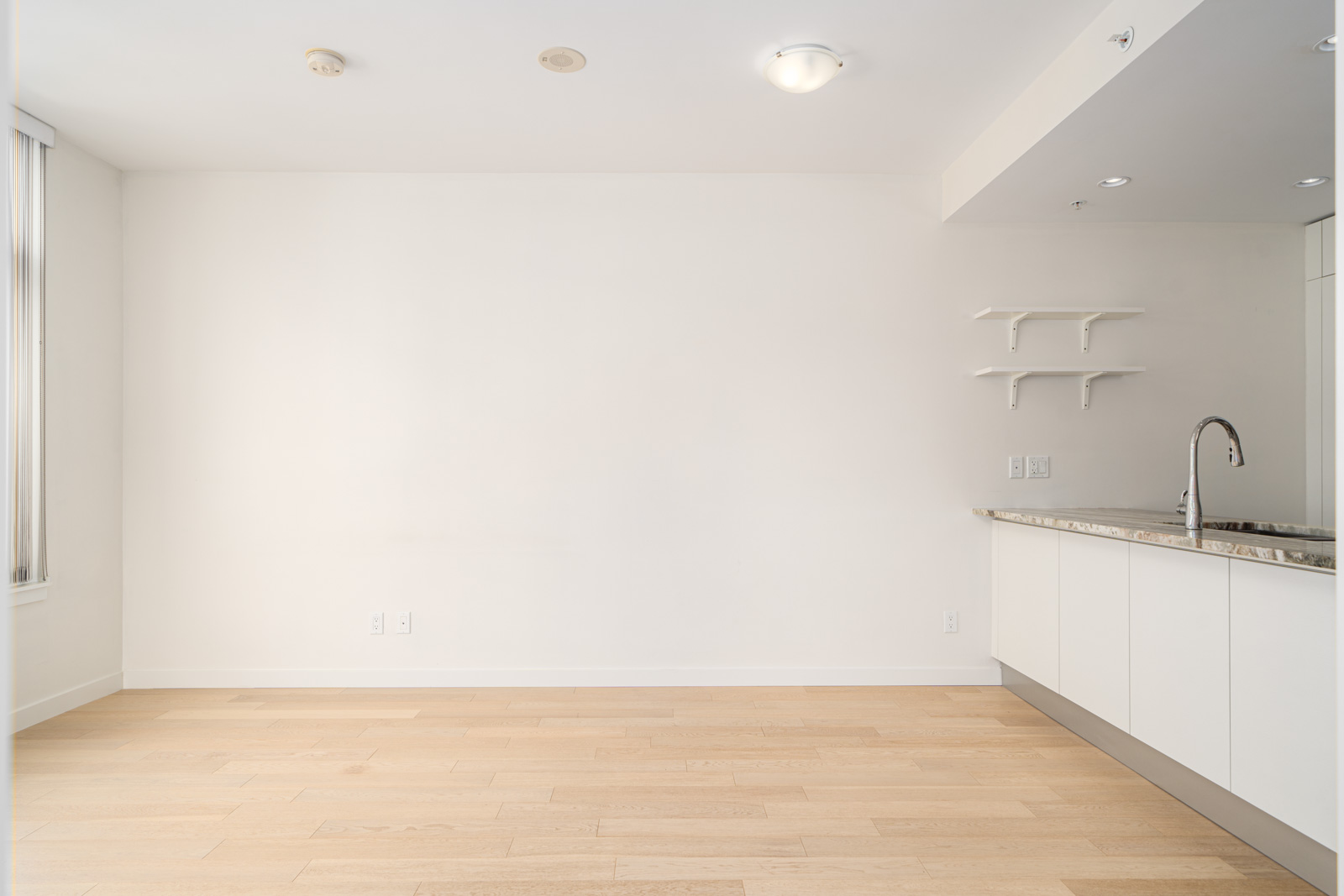 A modern, empty kitchen area with white walls, wooden flooring, a marble countertop, and white cabinetry. Two small wall shelves are mounted above the countertop near a stainless steel faucet.