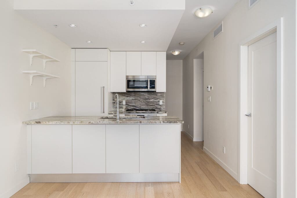 Modern kitchen with white cabinets, granite countertops, stainless steel appliances, and a small kitchen island. Light hardwood floors and minimalist shelving are present.