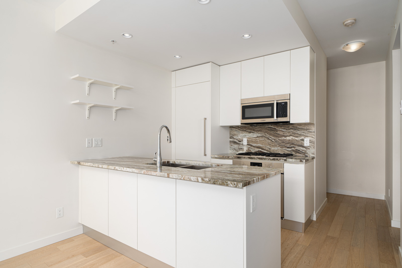 A modern kitchen featuring white cabinets, marble countertops, a built-in microwave, an oven, stainless steel sink with faucet, and two small wall-mounted shelves on the left. Light wood flooring.