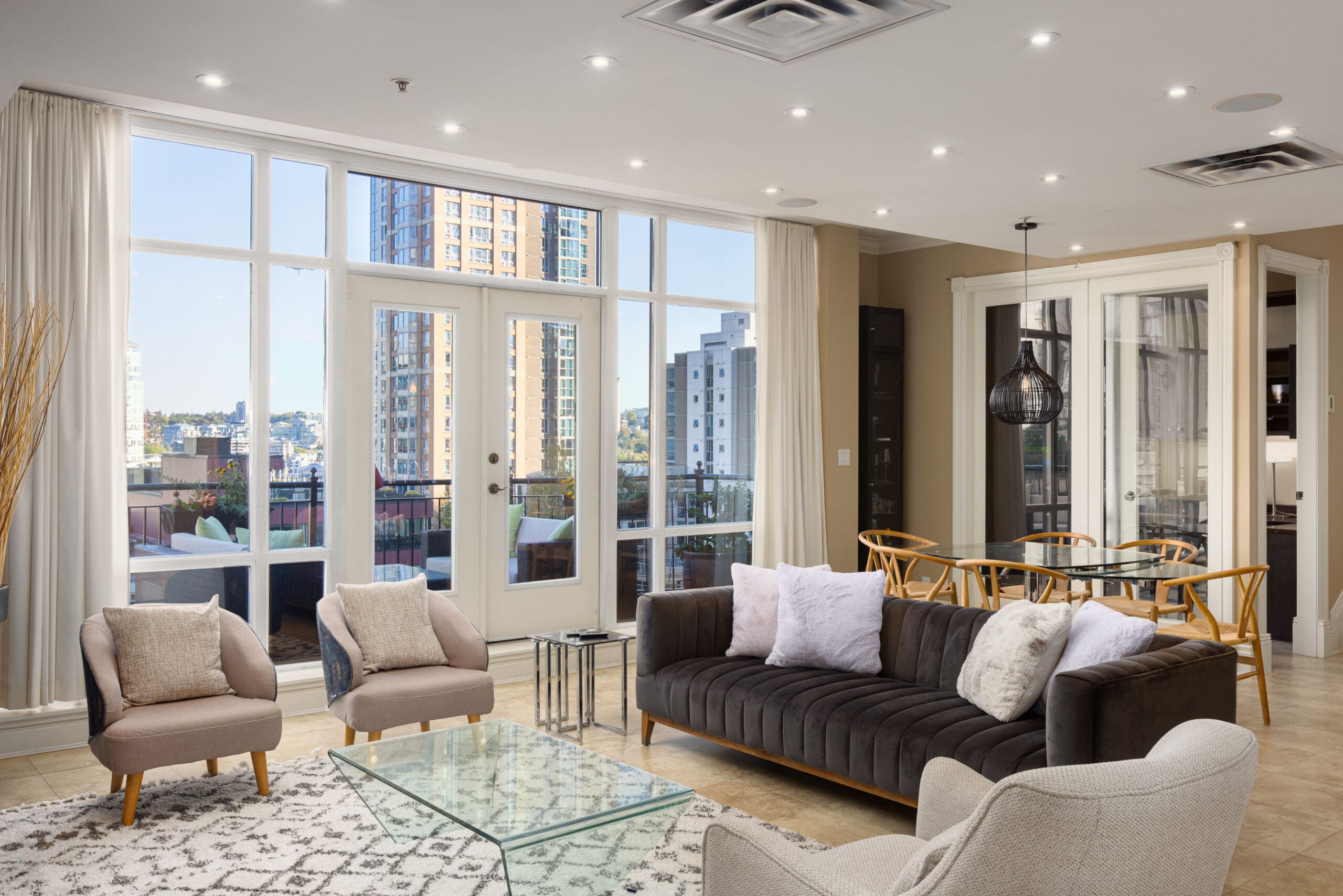 Modern living room with large windows, a dark sofa, light armchairs, a glass coffee table, and a view of city buildings. Dining area in the background has a black pendant light and glass table.
