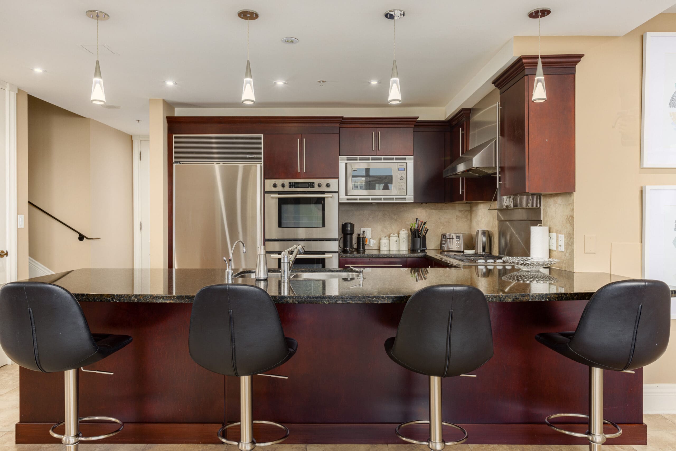 Modern kitchen with dark wood cabinets, stainless steel appliances, black countertop, and four black bar stools at the island. Pendant lights hang from the ceiling.