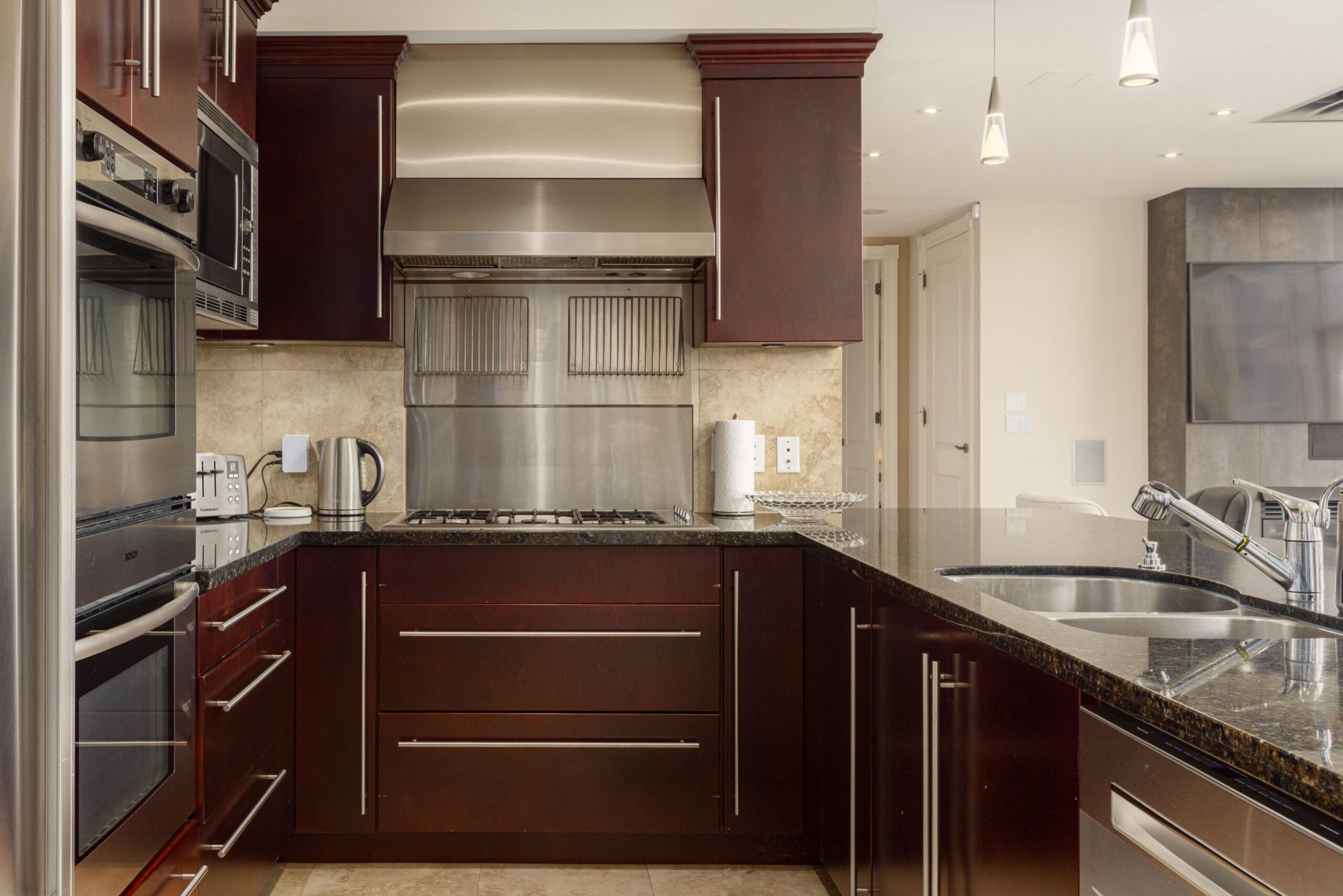 Modern kitchen with dark wood cabinets, stainless steel appliances, granite countertops, and a double basin sink. The backsplash features light beige tiles, and pendant lights hang above the island.