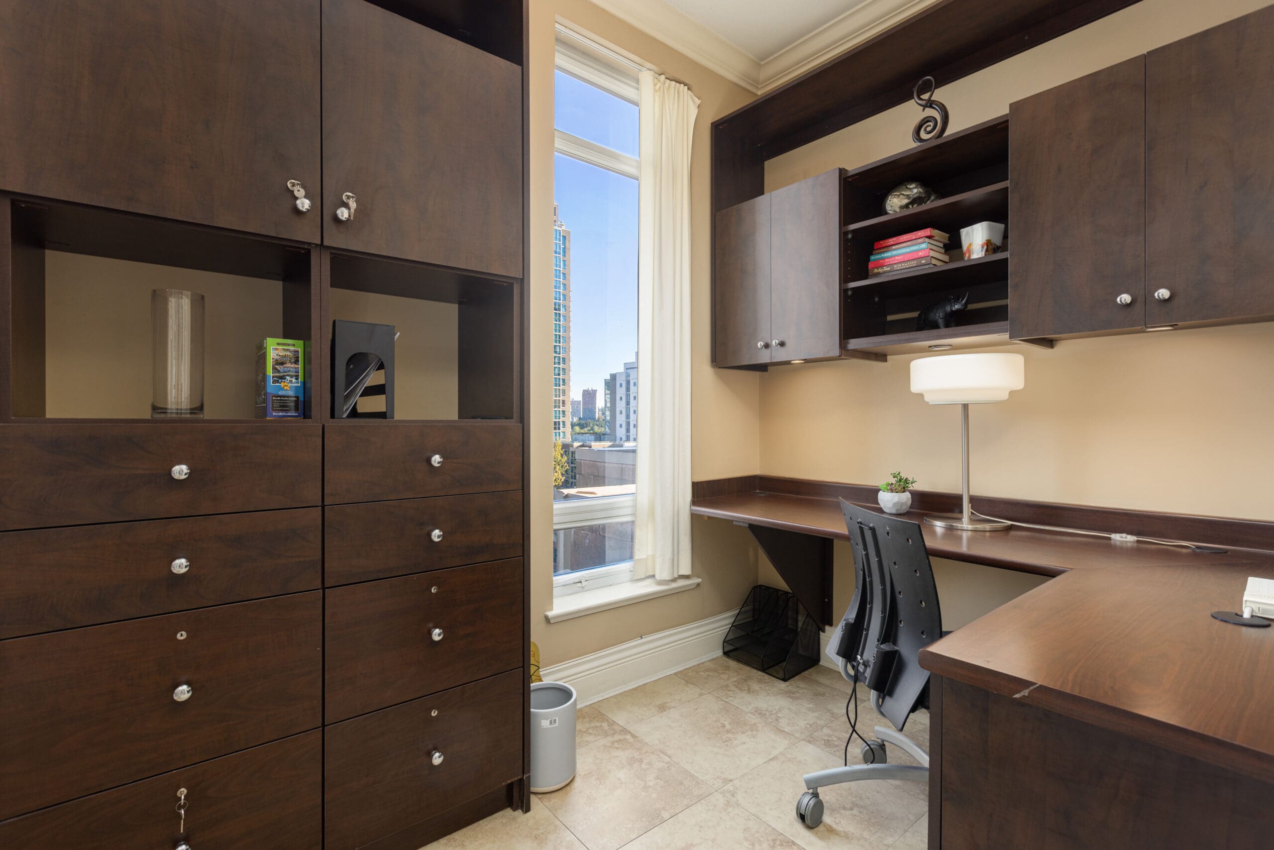 Modern home office with dark wood cabinetry, a desk, an ergonomic chair, books, a lamp, and a window showcasing a city view.