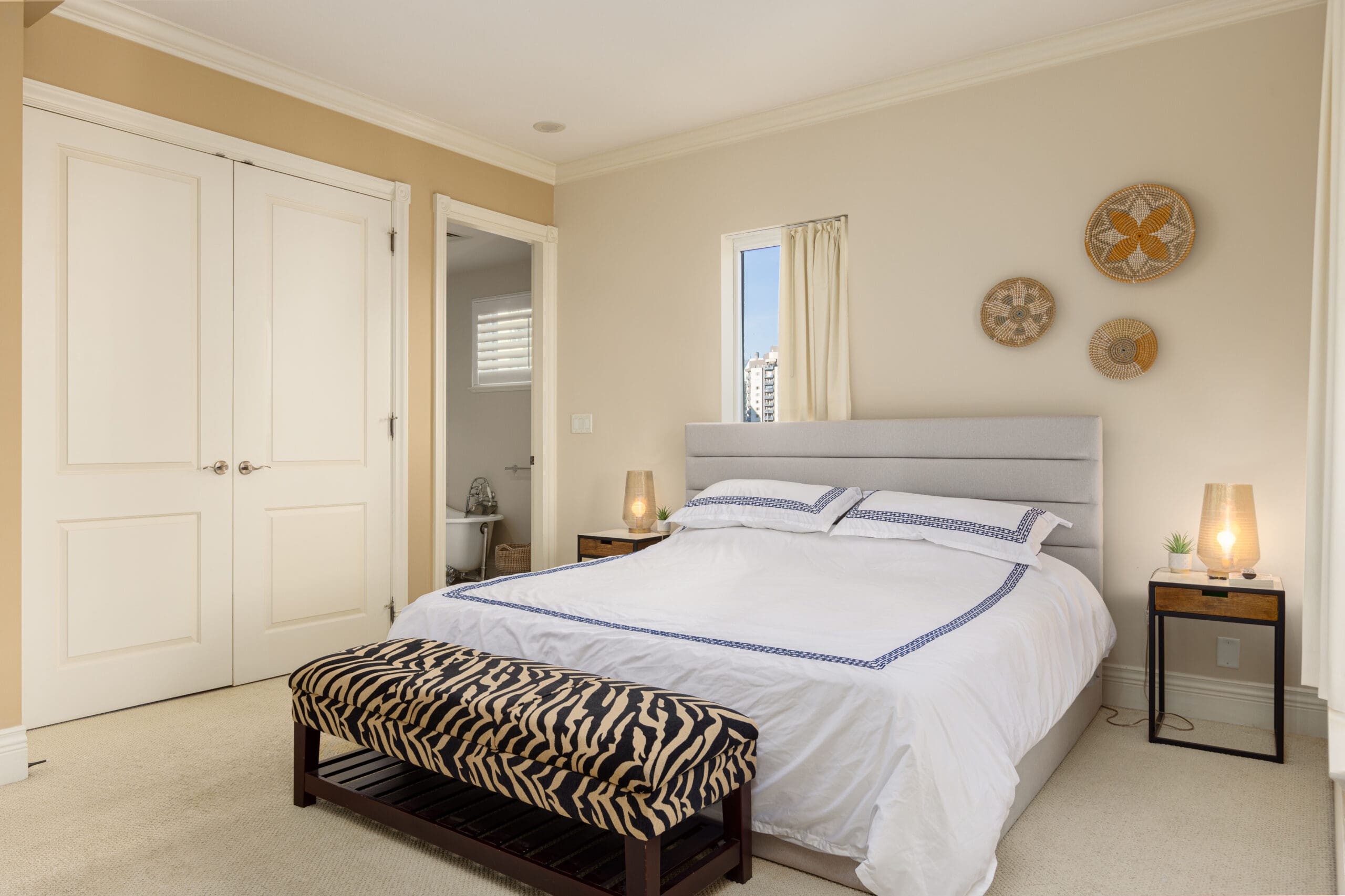 A bedroom with a double bed, a gray headboard, and white bedding. There is a zebra-print bench at the foot of the bed and two nightstands with lamps. A small window and wall decor are above the headboard.