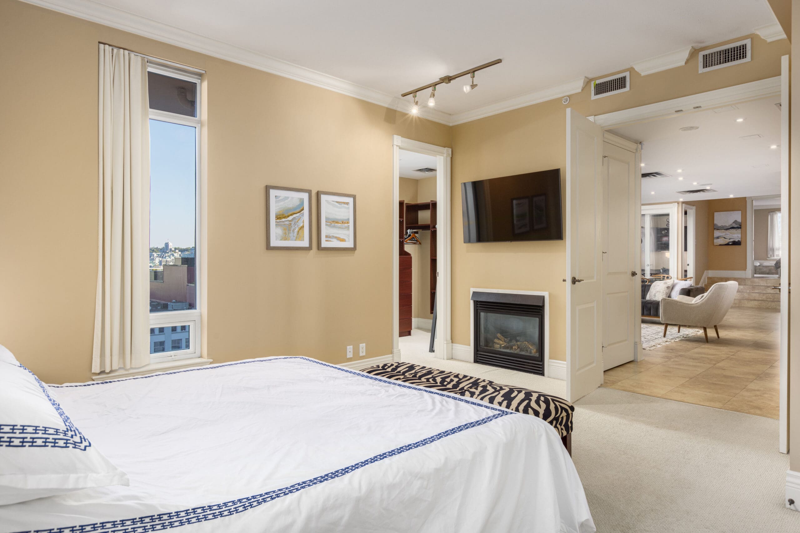 Bedroom with a view of a cityscape through a tall window, featuring a bed with white linen, a wall-mounted TV, a fireplace, and connected to a spacious open-plan living area.