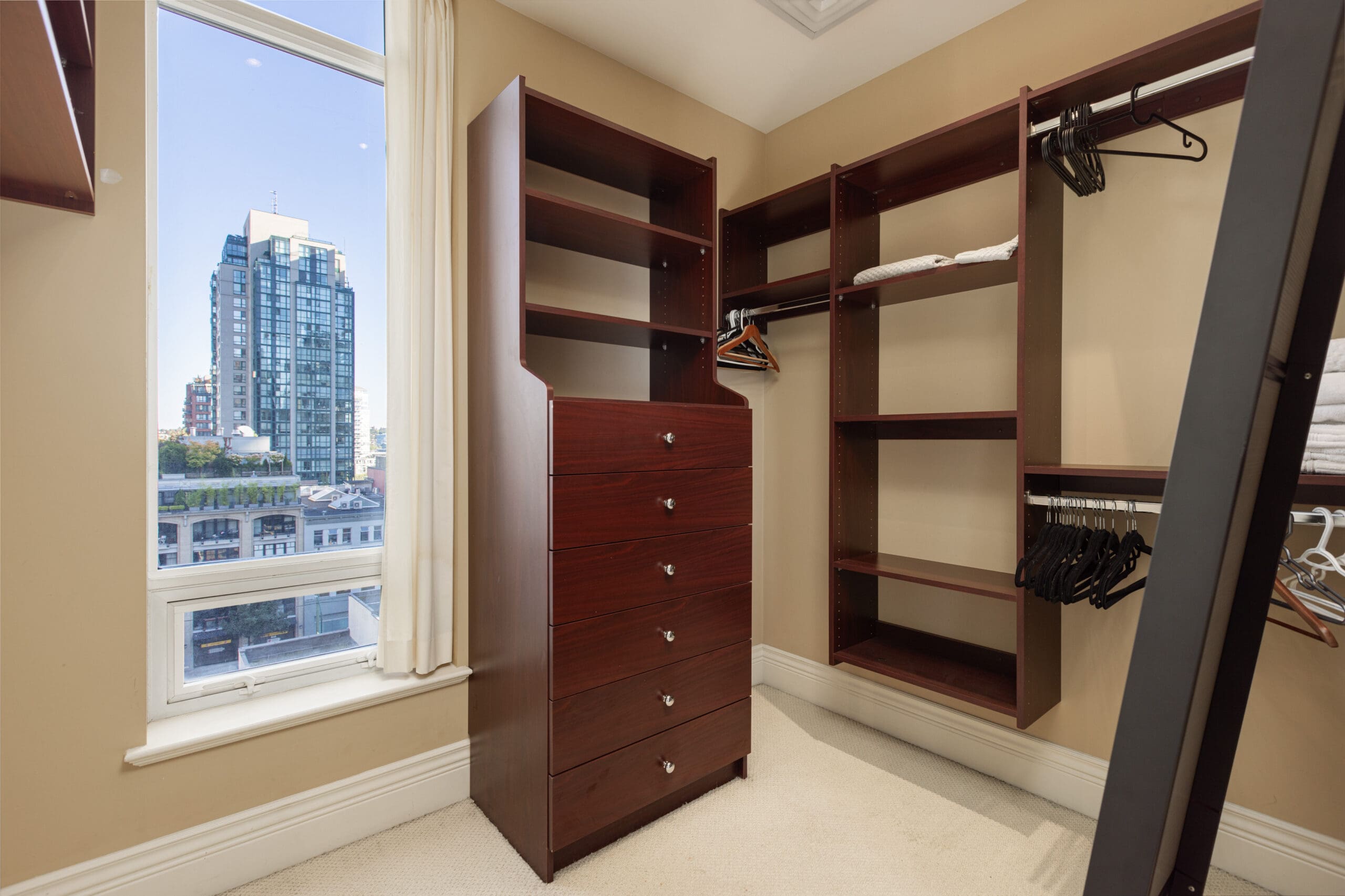 A walk-in closet with wooden shelves and drawers, some hangers and clothes, and a large window offering a city view.