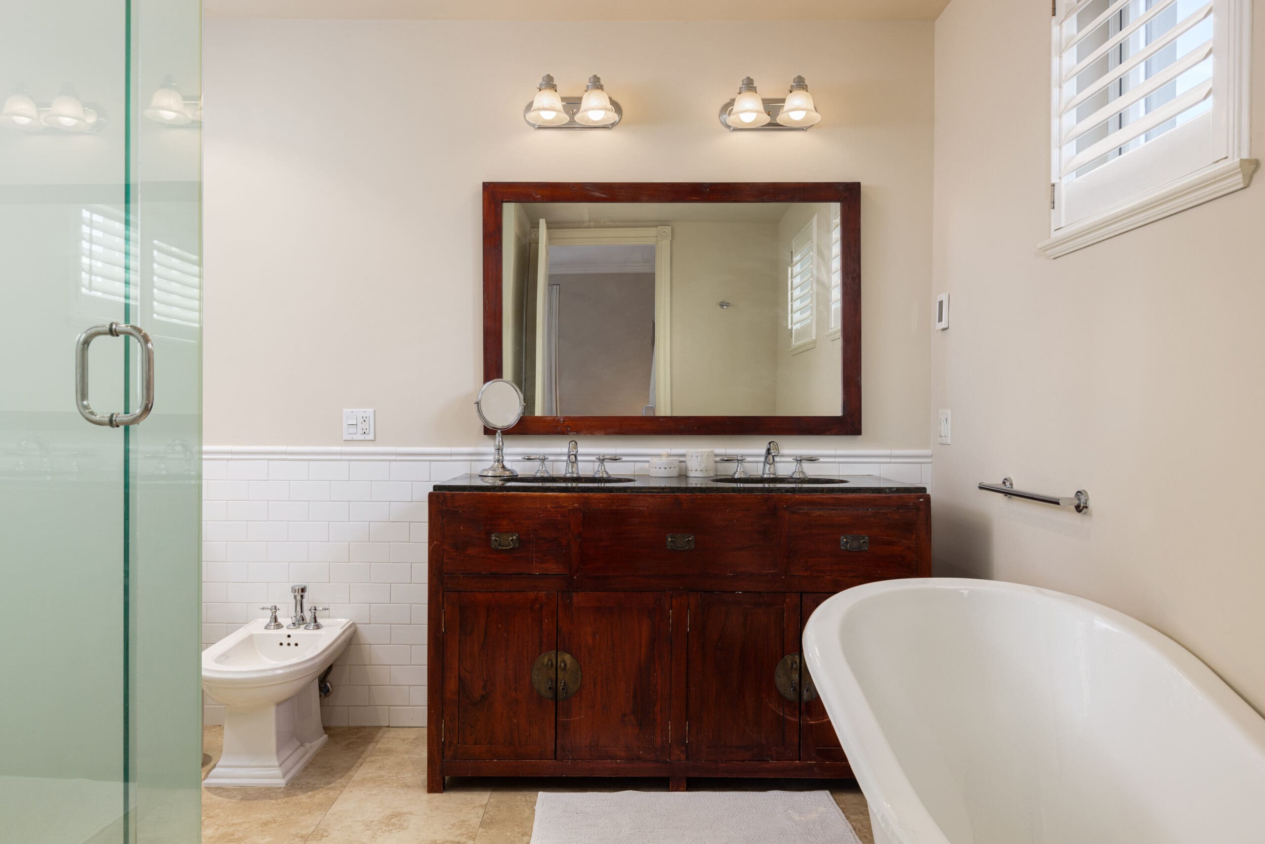 A bathroom with a wooden vanity and double sinks, a large mirror, a bidet, and a freestanding bathtub. There is a glass door shower and wall-mounted light fixtures above the mirror.