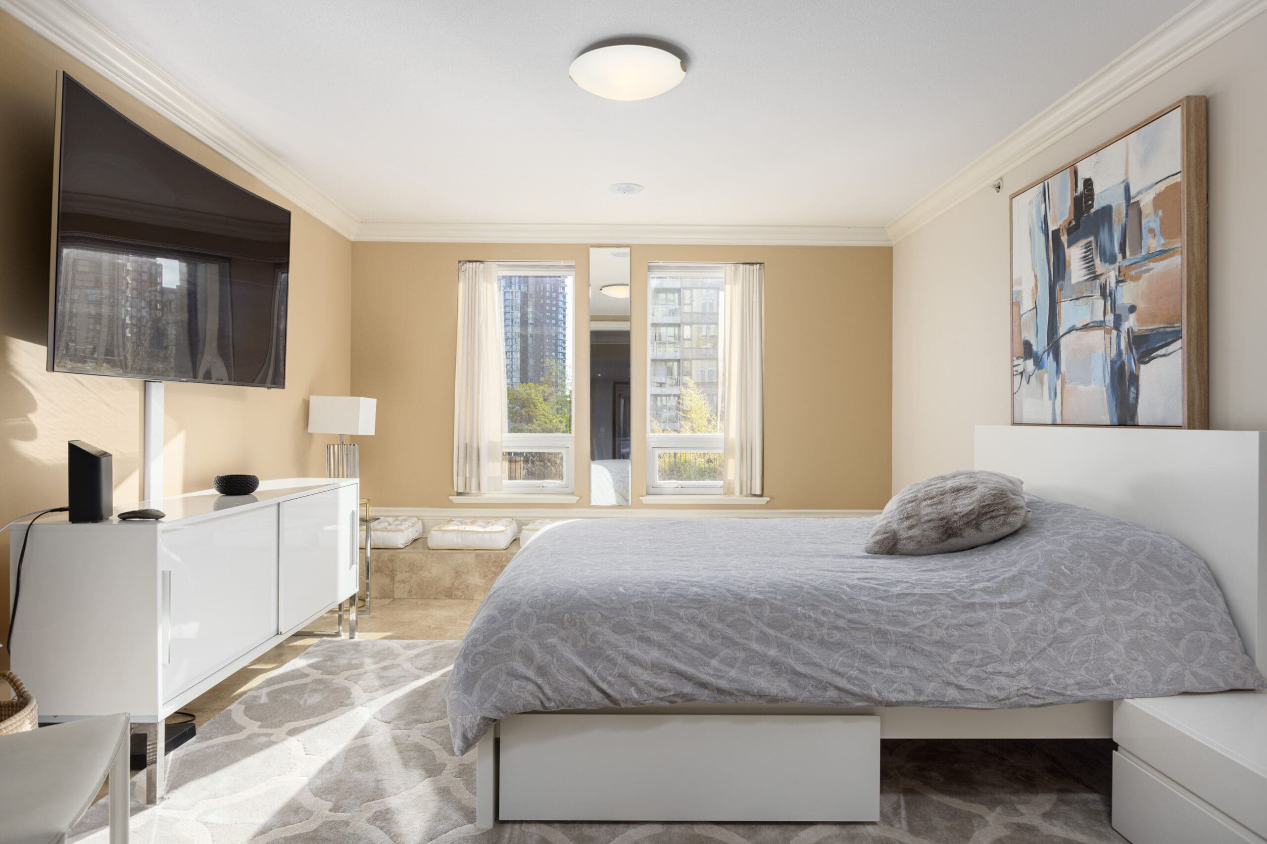 A modern bedroom with a large bed, abstract artwork above the headboard, a wall-mounted TV, two windows providing natural light, and neutral-colored furnishings.