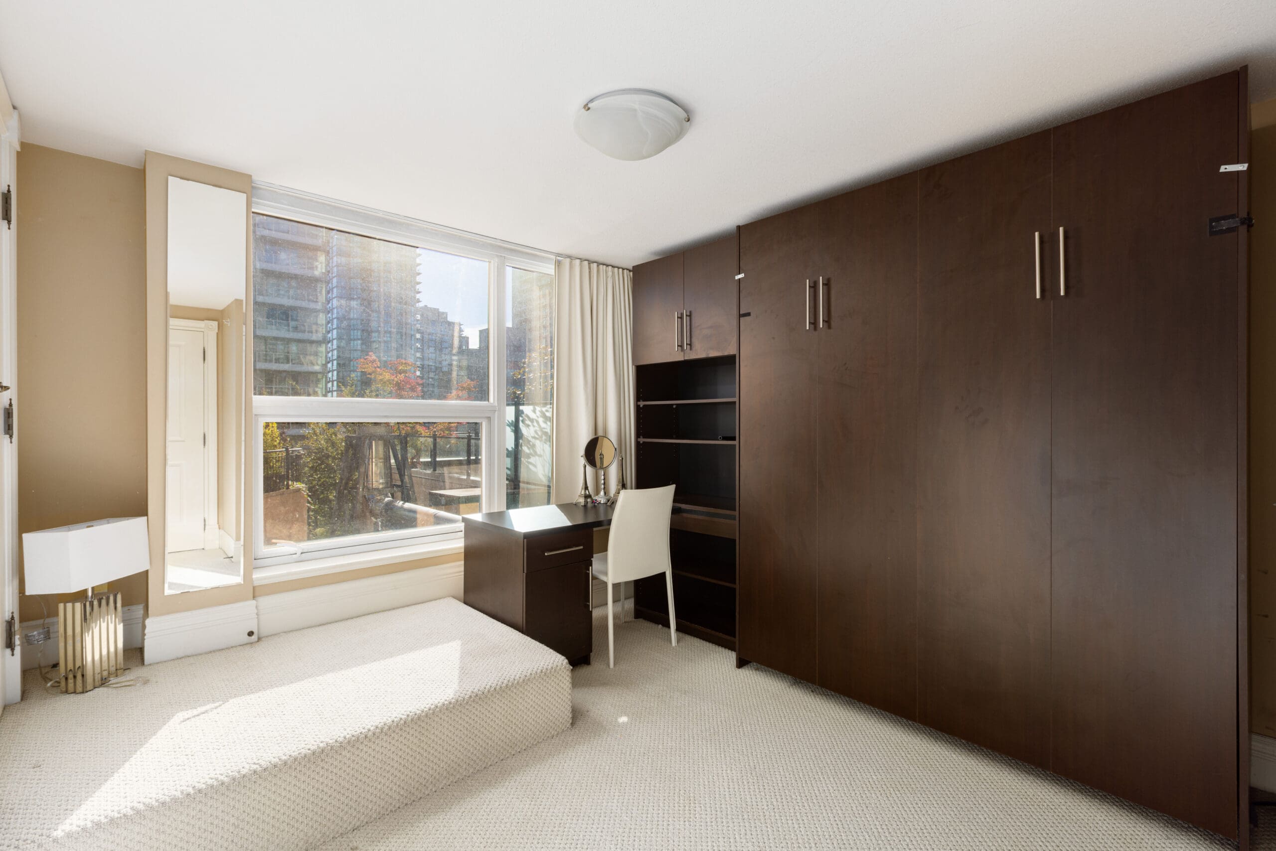 A bedroom with a large window, built-in dark wood closet, desk, chair, and a small vanity. Floors are carpeted in light color. A floor lamp and a mirror are by the window.