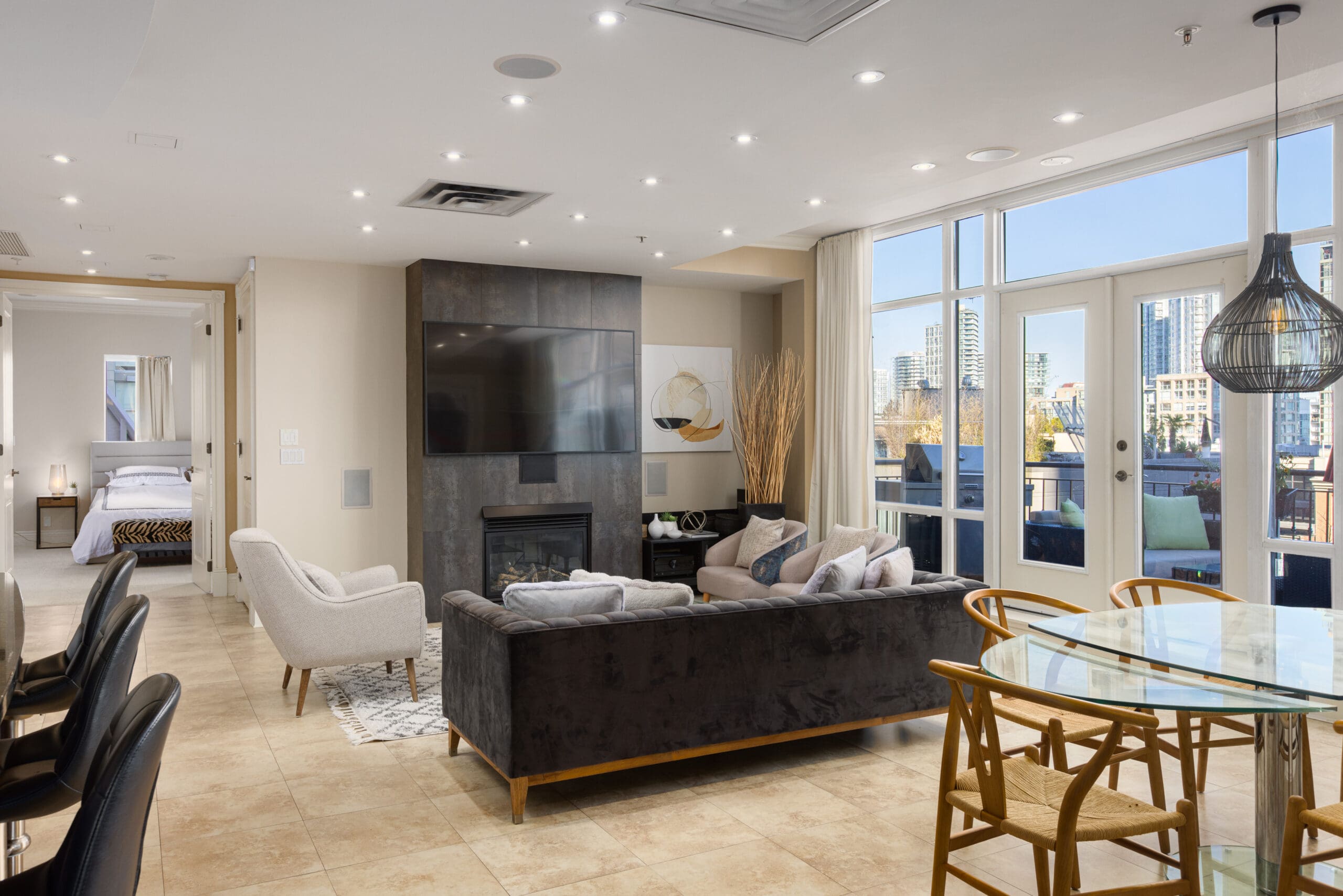 A modern open-concept living space featuring a living room with a wall-mounted TV, a dining area with a glass table, and a view of a cityscape through large windows. A bedroom is visible in the background.