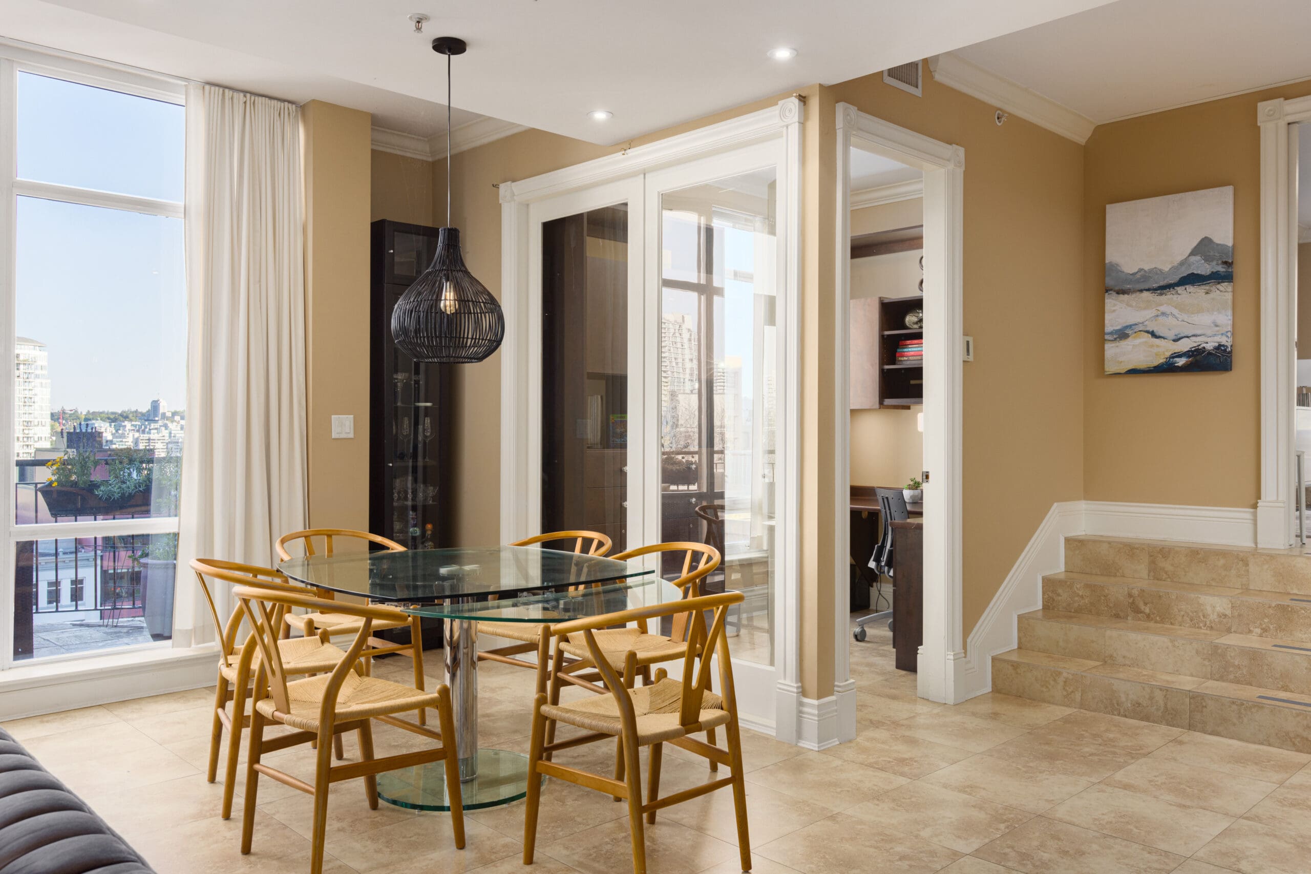 A modern dining area with a glass table, wooden chairs, large windows, and a black pendant light. A small office nook is visible through a glass door next to stairs leading to another level.
