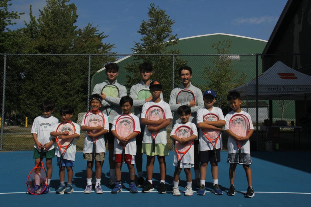 A group of young boys holding tennis rackets pose with three adult men on a blue tennis court, with a green building and trees in the background.