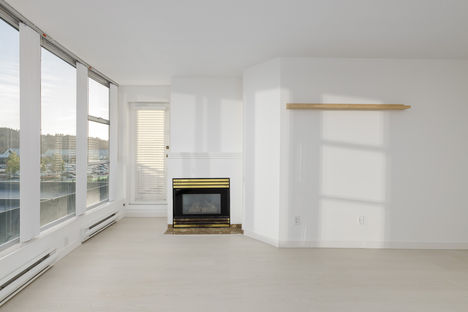 Empty living room with large windows, a fireplace, and a wooden floating shelf on a white wall. Hardwood floors and natural light fill the space.
