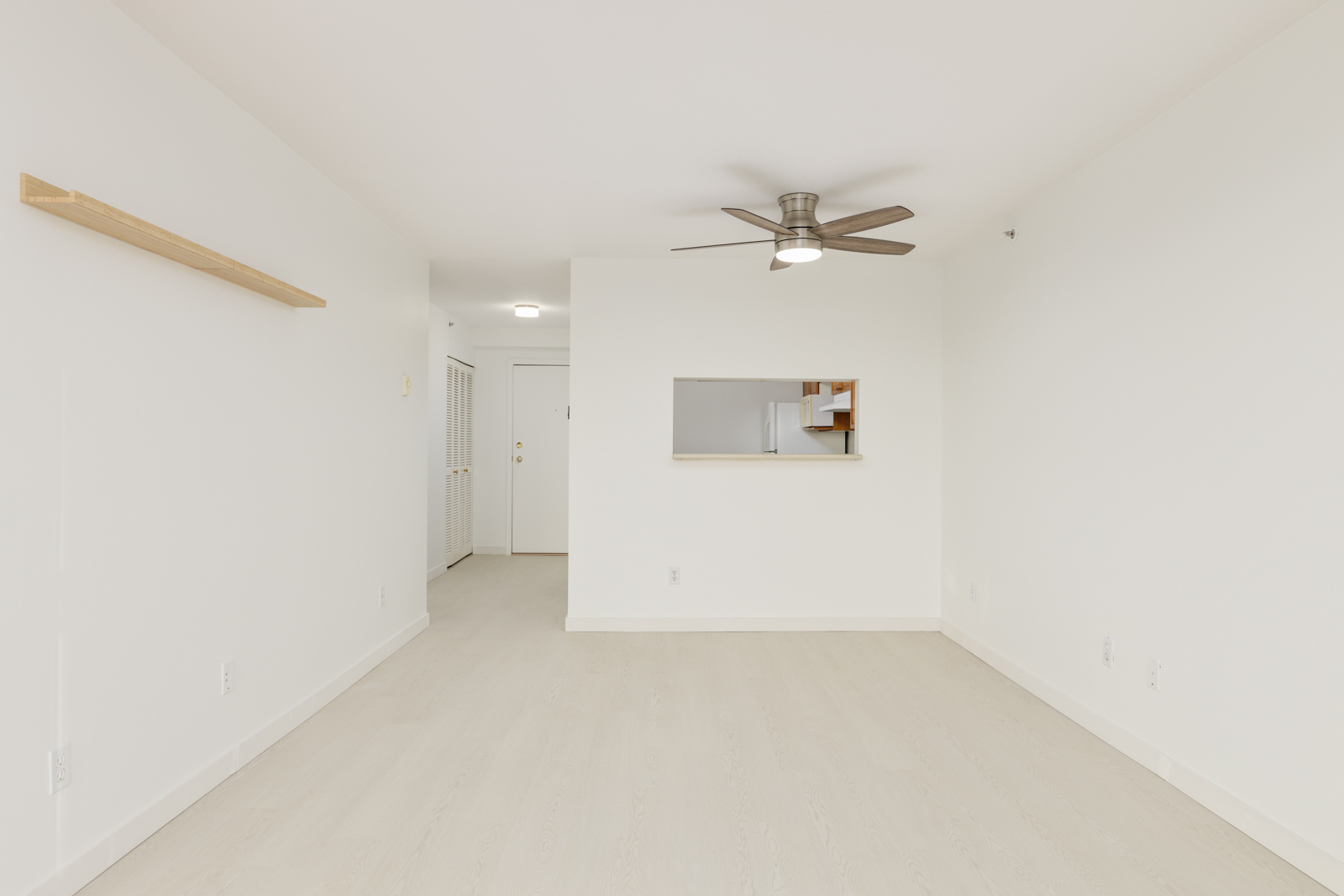 Empty white room with light wood flooring, a ceiling fan, a wall shelf, and a rectangular opening in the wall showing another room.