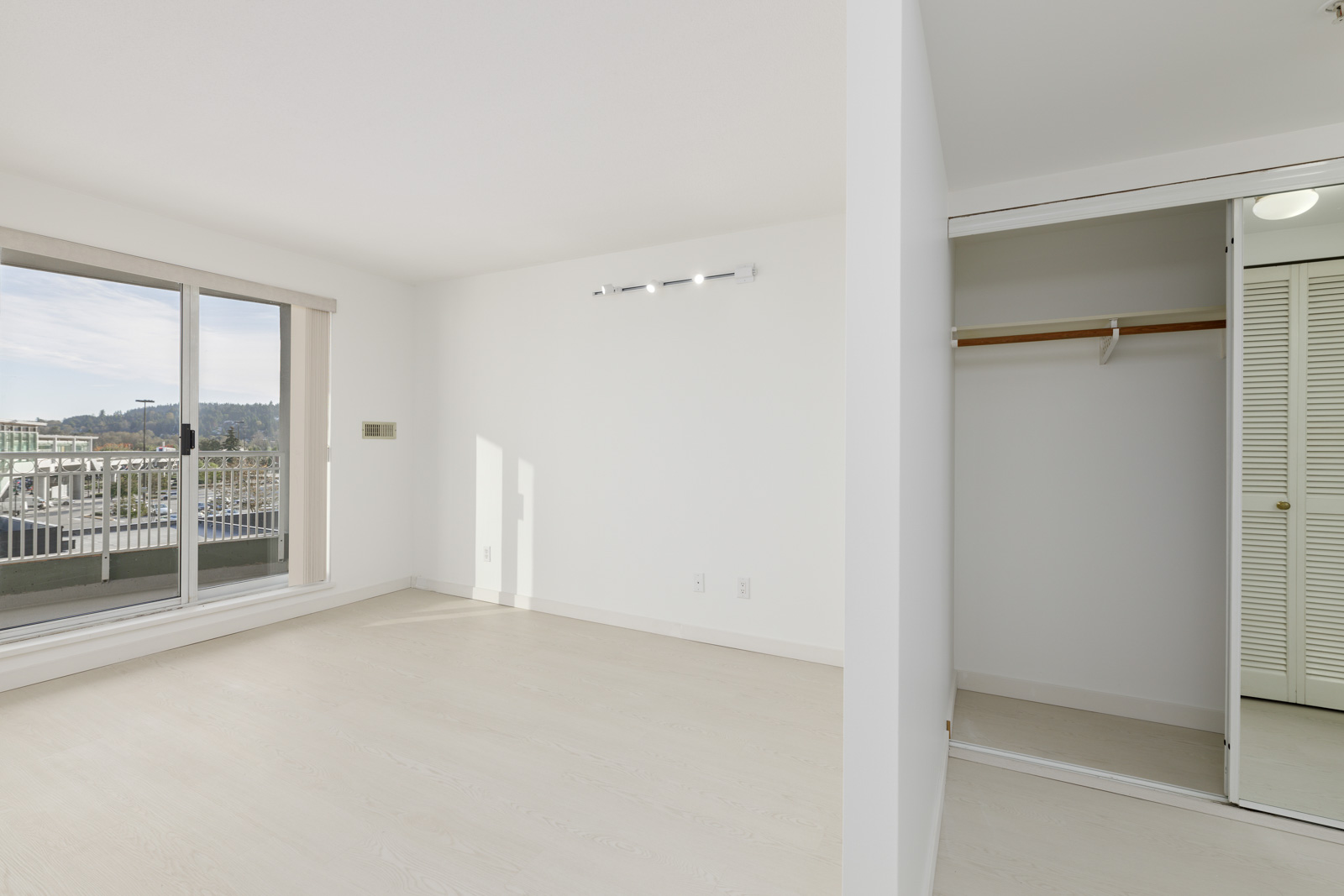 Empty, well-lit room with light wood flooring, sliding closet doors, and a window view of a balcony and cityscape.
