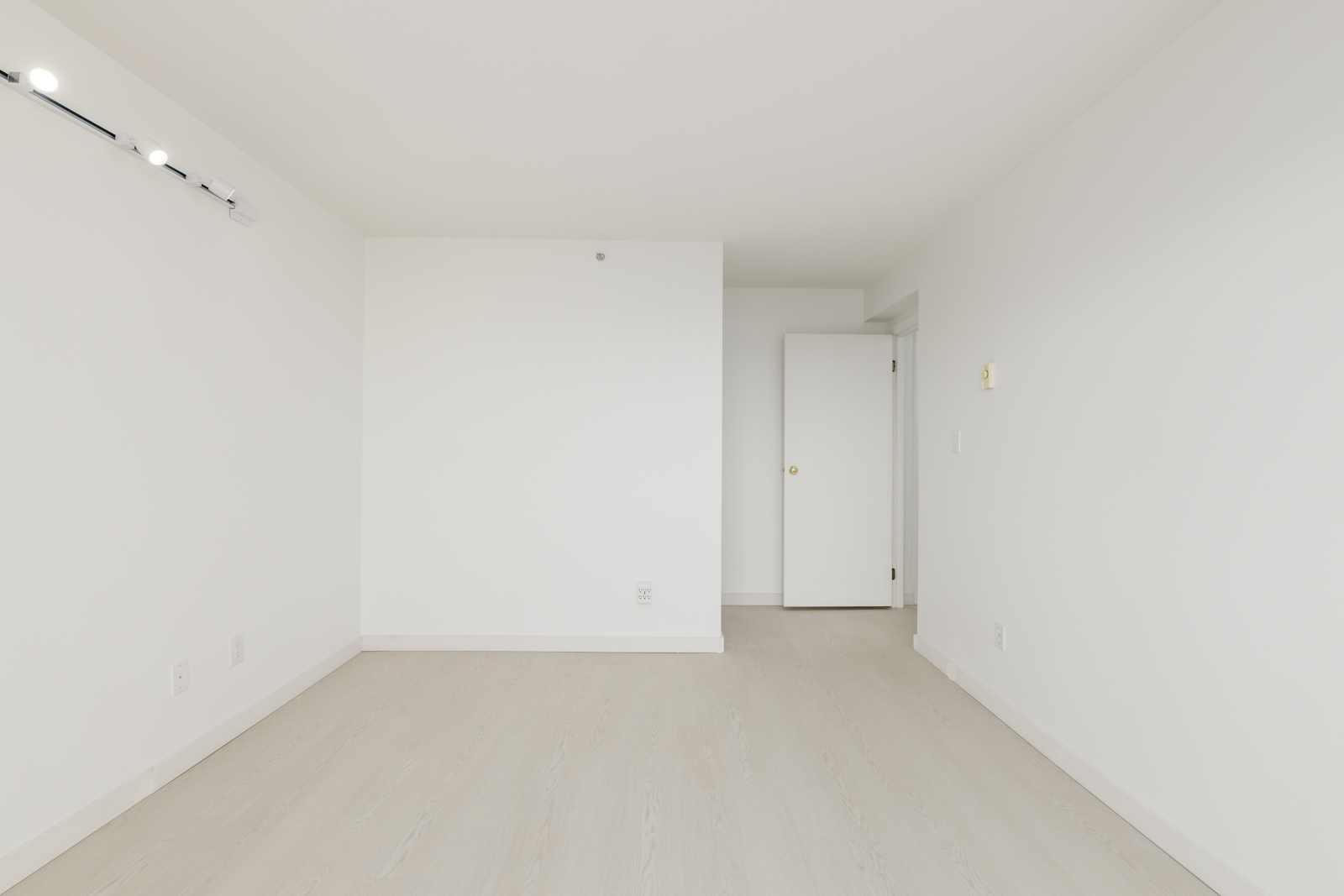 A vacant, unfurnished room with white walls, light wood flooring, a closed door, and a track lighting fixture on the ceiling.