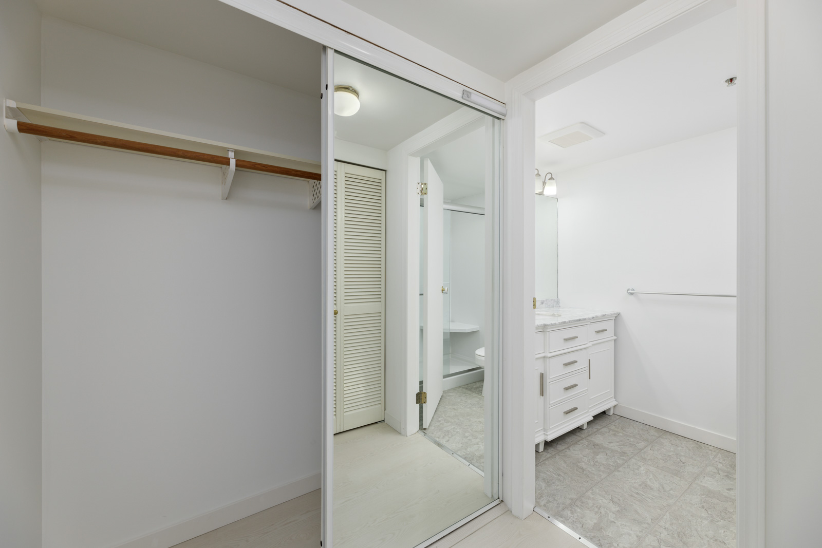 White walk-in closet with sliding mirrored doors, leading to a bathroom with a white vanity and gray tile flooring.