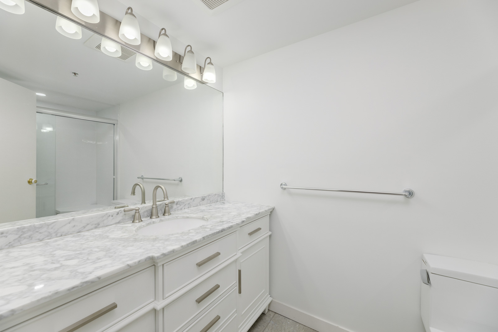 A modern bathroom with a white double-sink vanity, marble countertop, large mirror, and overhead lights. A towel bar and a closed toilet seat are visible.