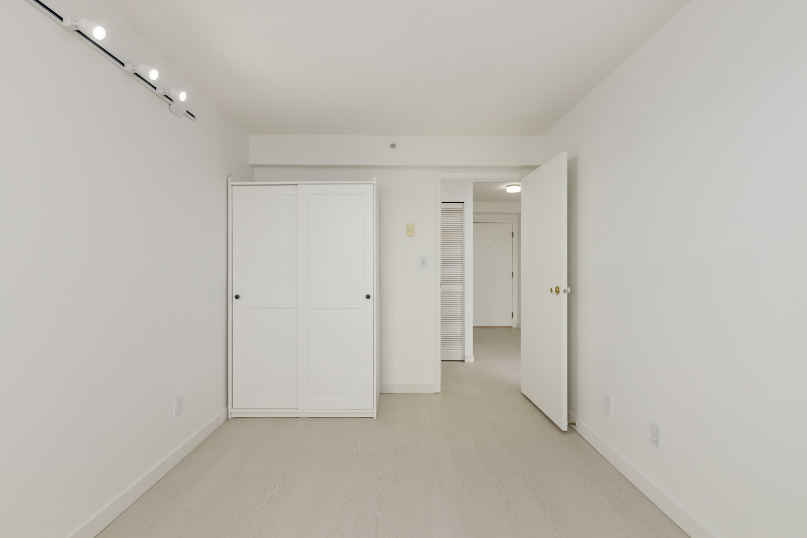 Empty room with white walls, light wood flooring, a white wardrobe, and an open door leading to a hallway. Ceiling track lights are visible.