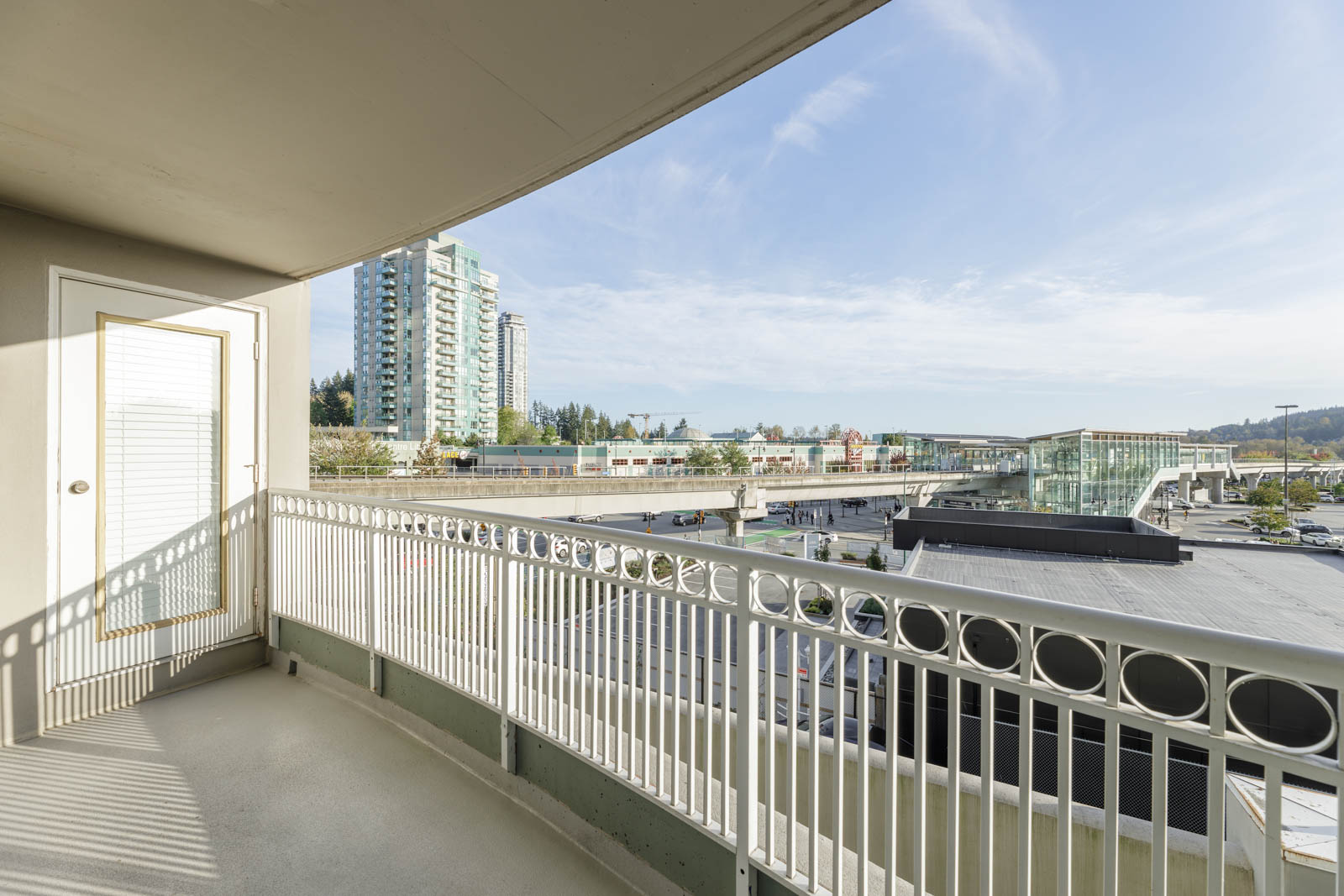 A balcony with a metal railing overlooks a cityscape with tall buildings, a train station, and a clear sky.