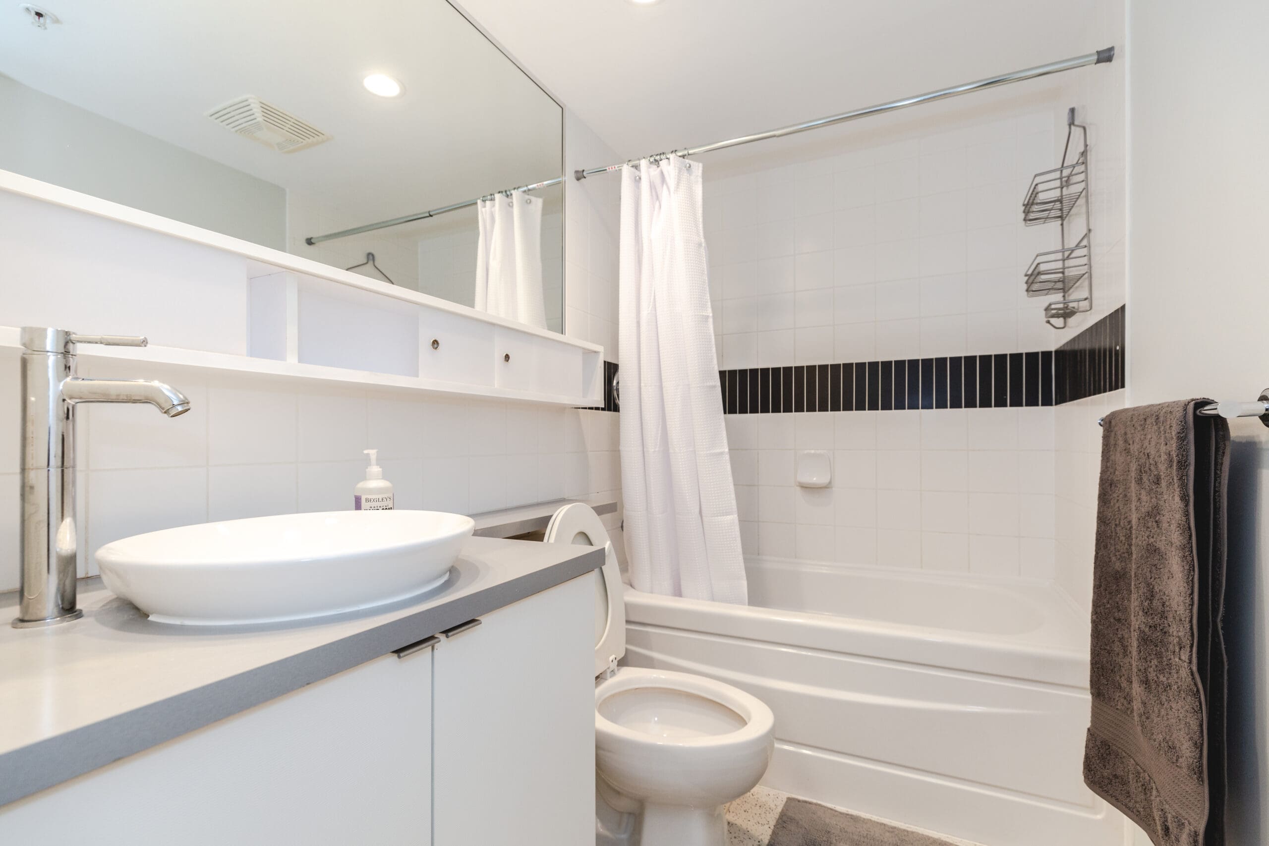 Modern bathroom with white fixtures, including a sink, toilet, and bathtub with a shower curtain. A mirror runs along one wall above a countertop and towel rack.